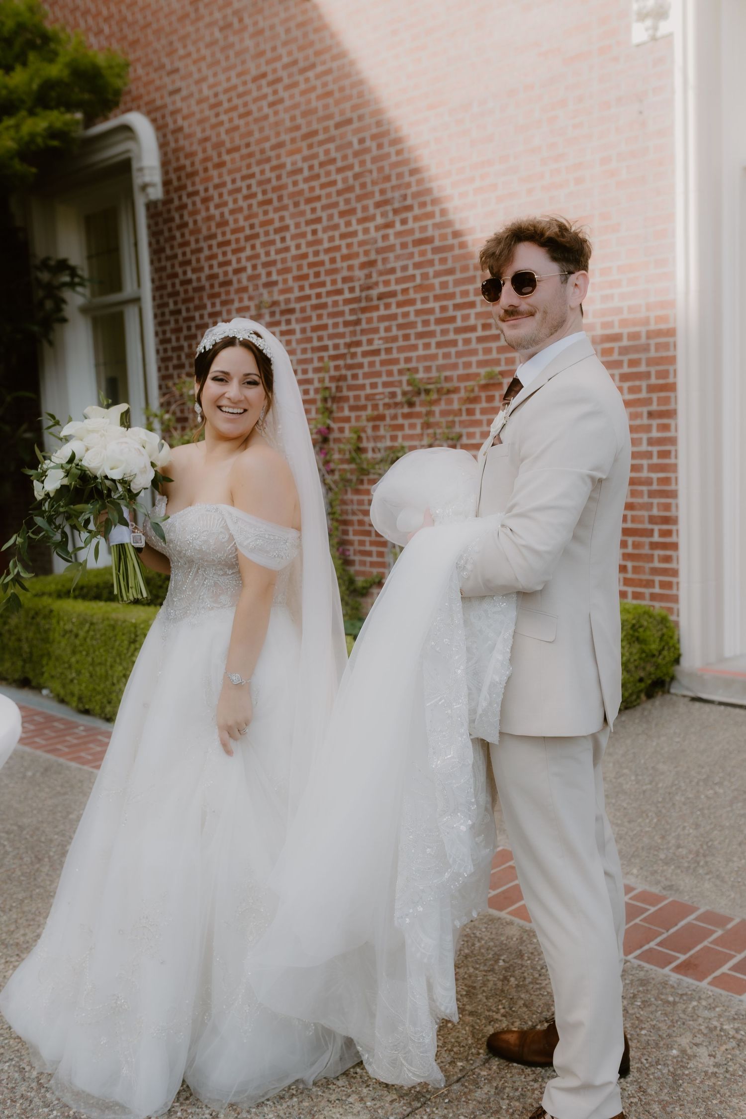Smiling bride in off-shoulder gown stands with groom in tan suit holding her wedding veil.