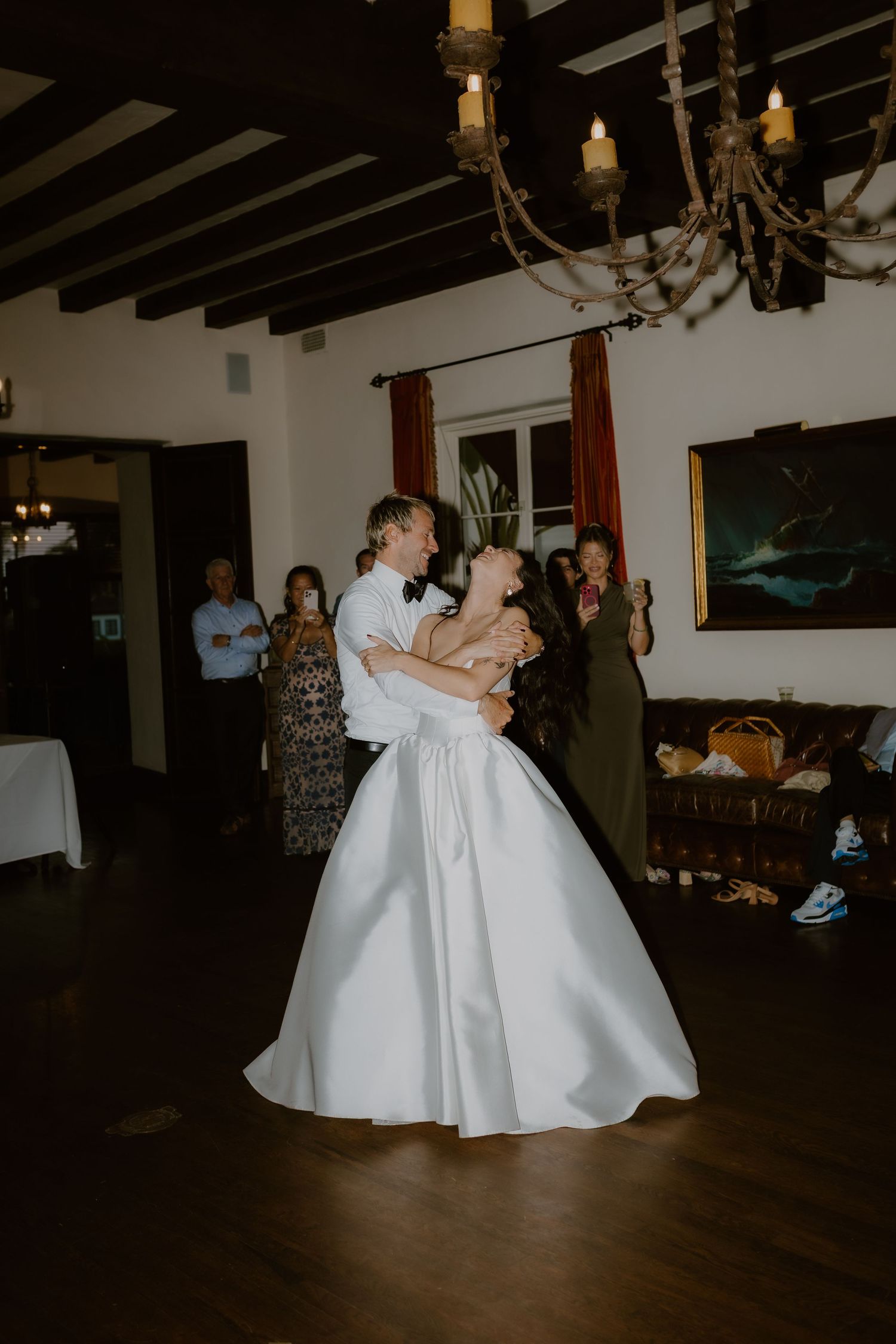 Bride and groom sharing first dance in elegant rustic reception hall with chandelier lighting.