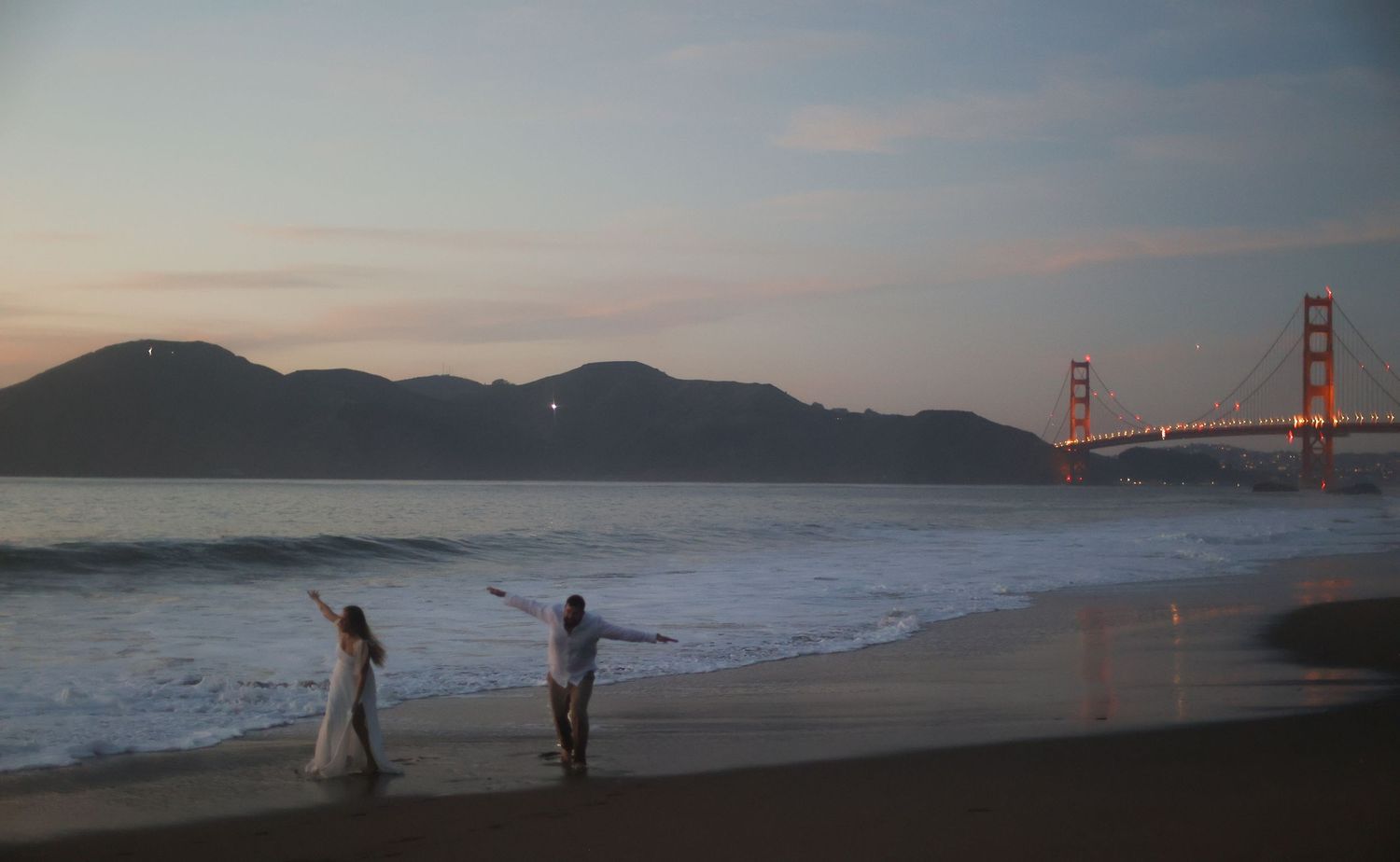 Newlyweds celebrating on a sandy beach at dusk with the iconic Golden Gate Bridge glowing behind them.