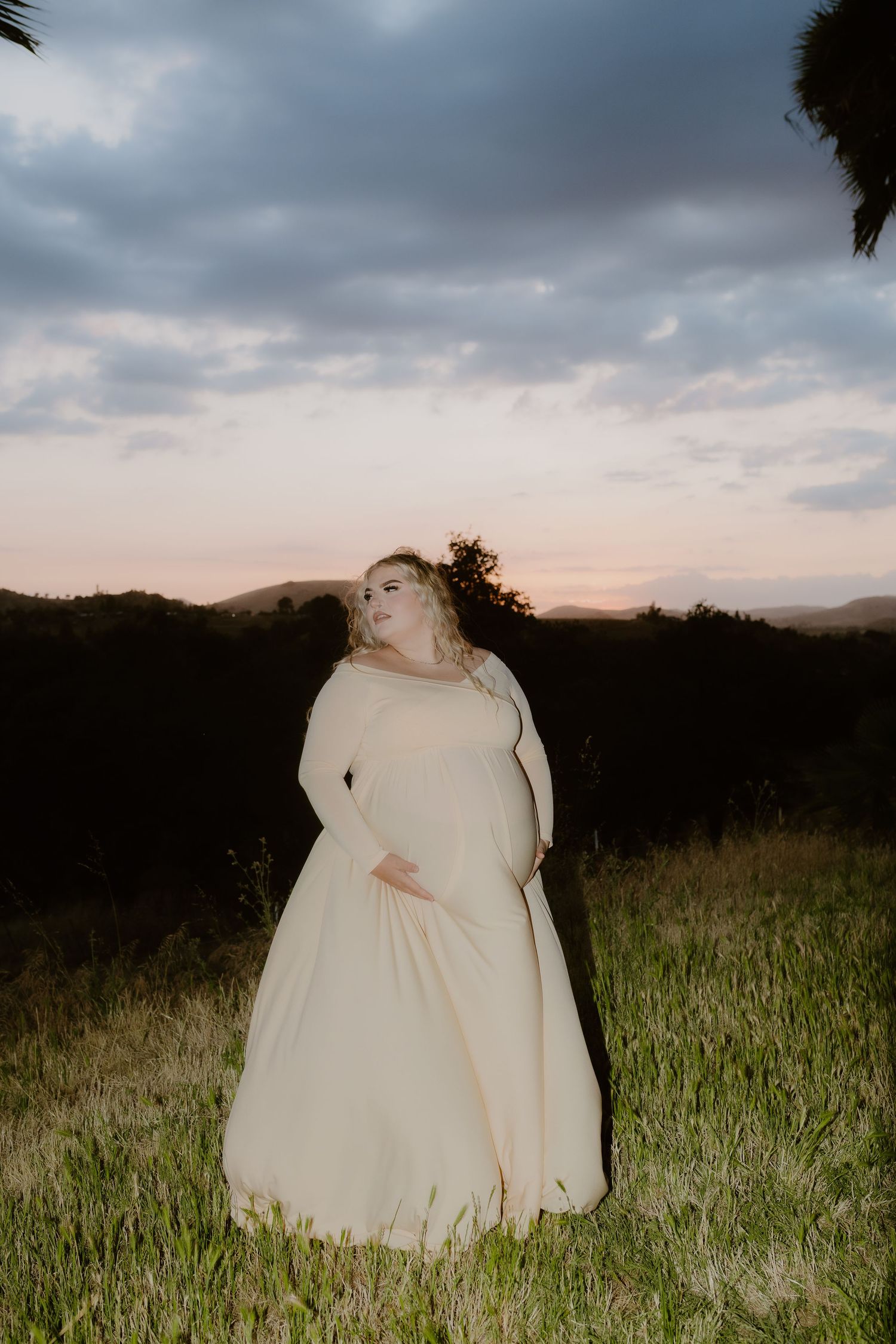 Pregnant woman in cream gown standing in a field at sunset with rolling hills behind her. maternity session, editorial, direct flash