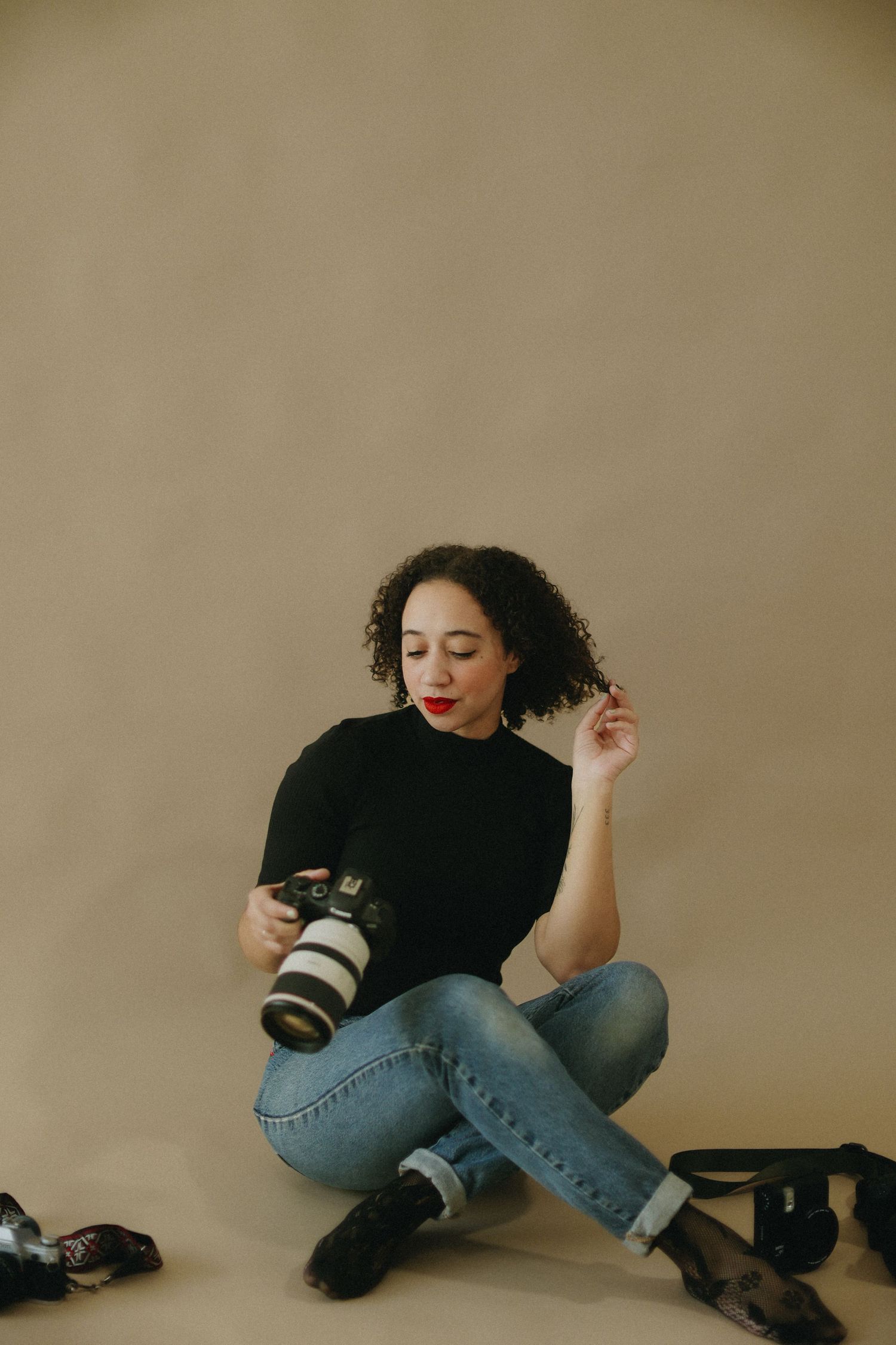 Curly-haired woman in black top holds Canon camera while sitting on beige studio floor. photographer