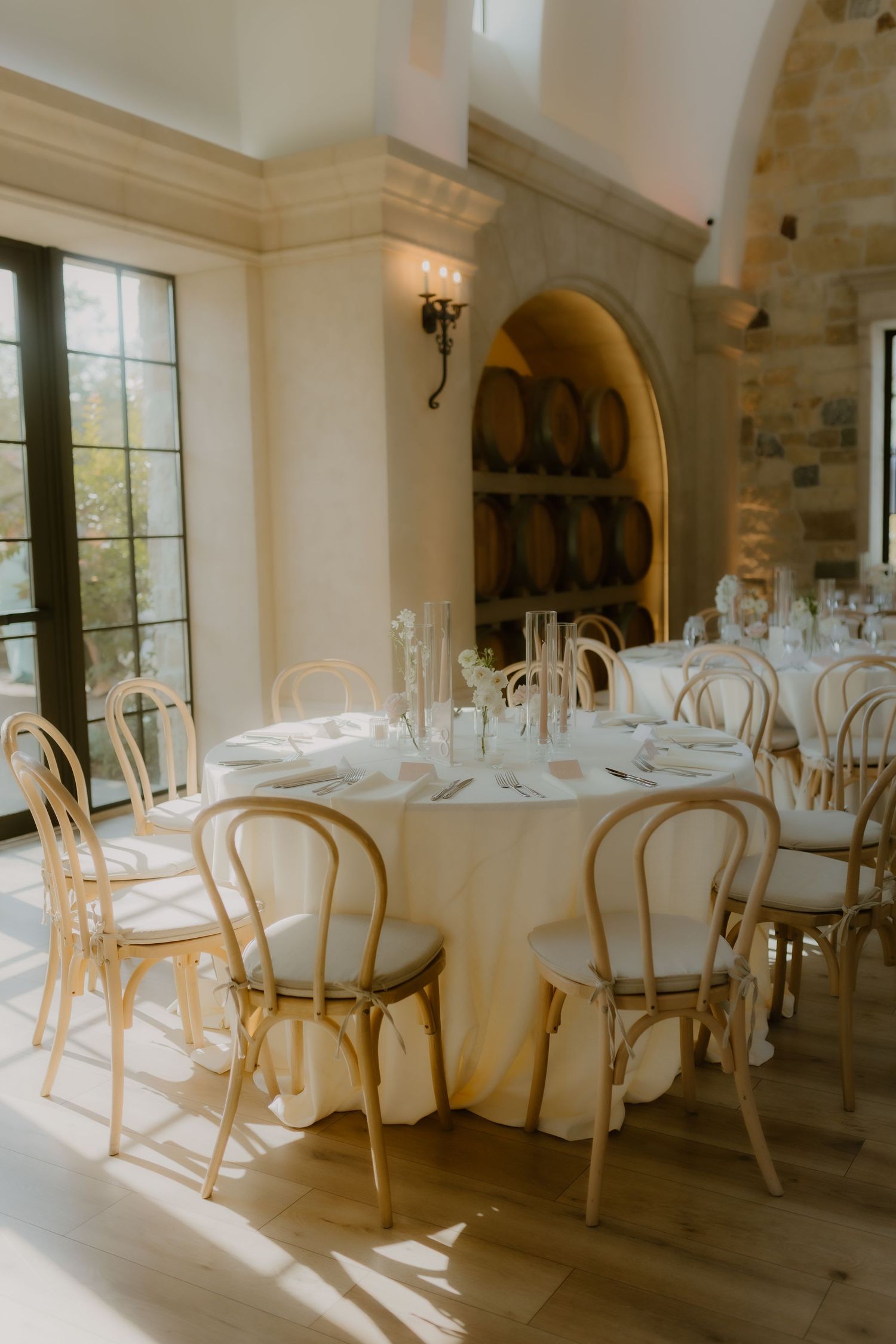 Sunlit wedding reception room with wooden bentwood chairs, white tablecloths, and wine barrel alcove.