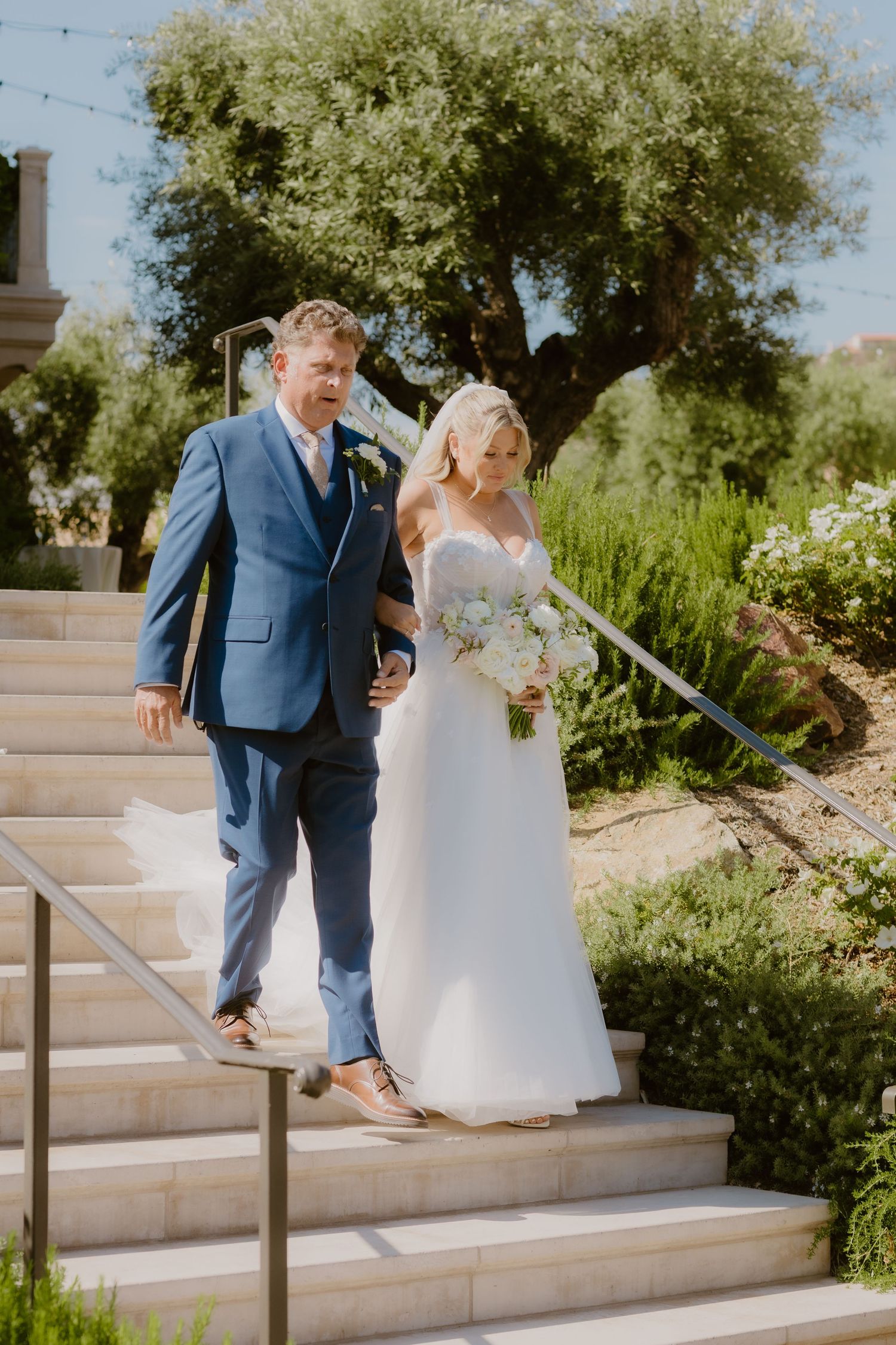 Bride and groom walking down outdoor stone steps at a sunny wedding venue surrounded by lush greenery.