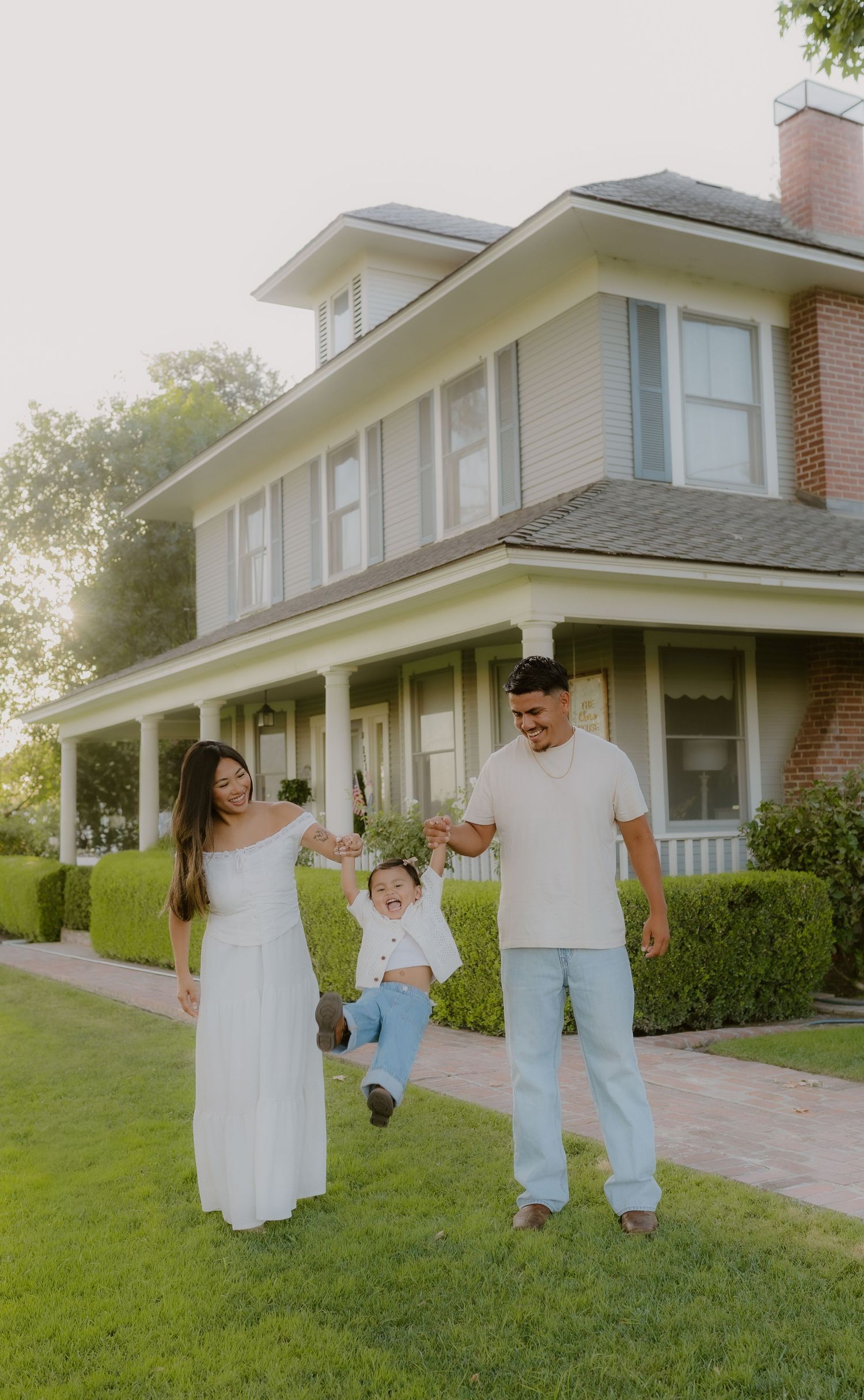 Smiling family lifting toddler between them on green lawn outside classic American suburban home.