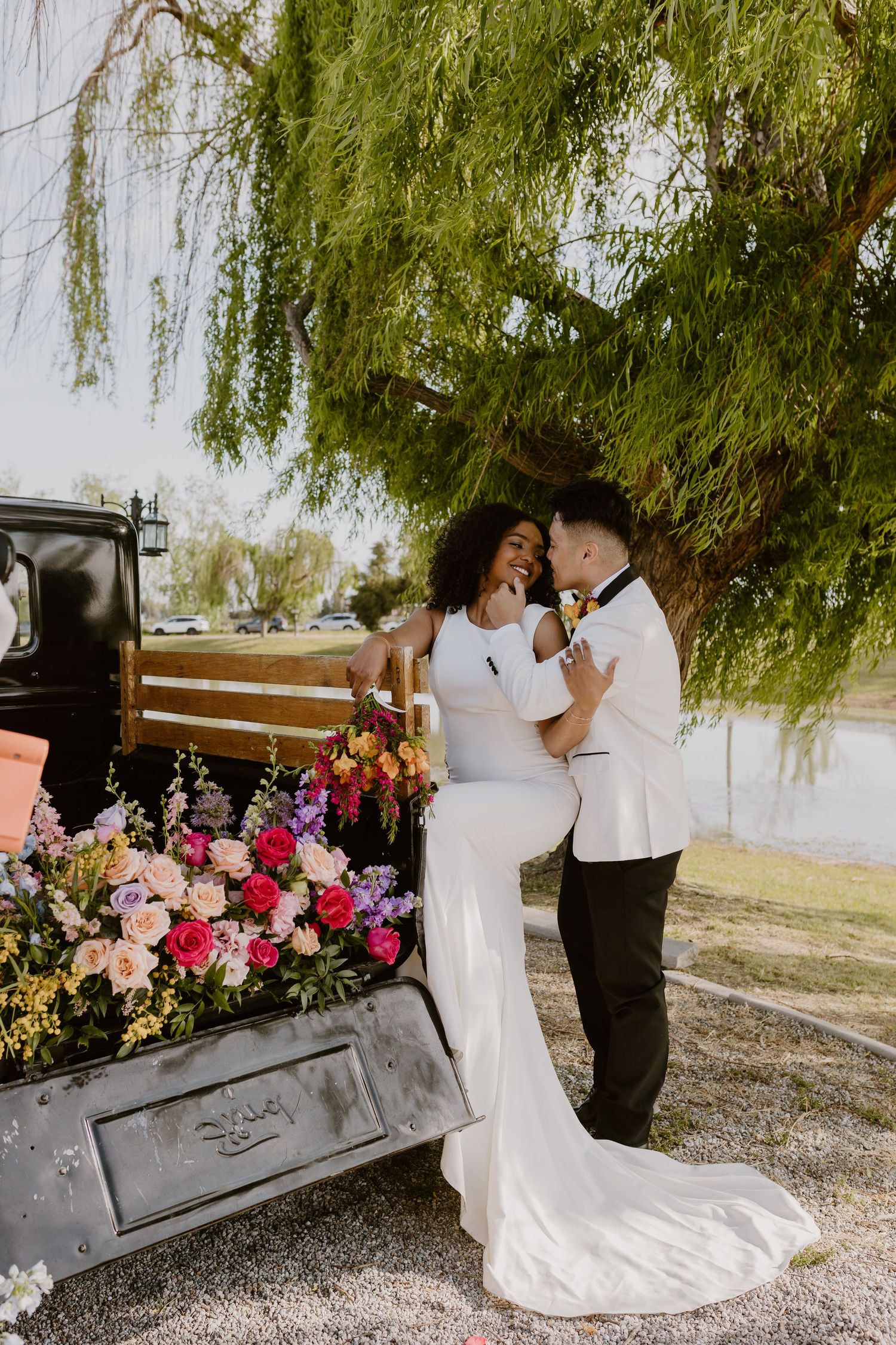 Bride and groom share a romantic moment beside a vintage truck adorned with colorful floral arrangements under a willow tree.