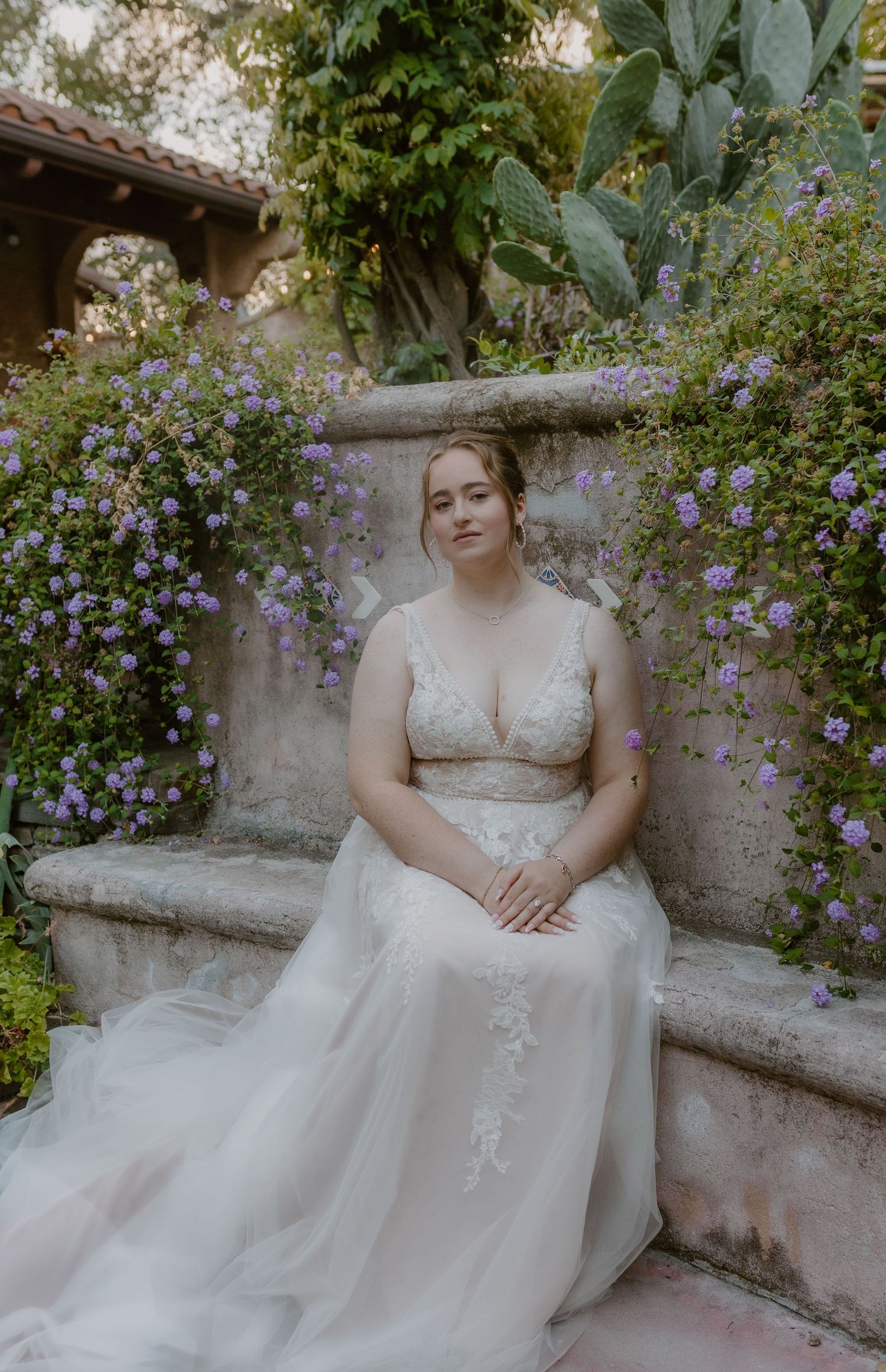 Bride in lace tulle gown seated among purple flowering garden walls.
