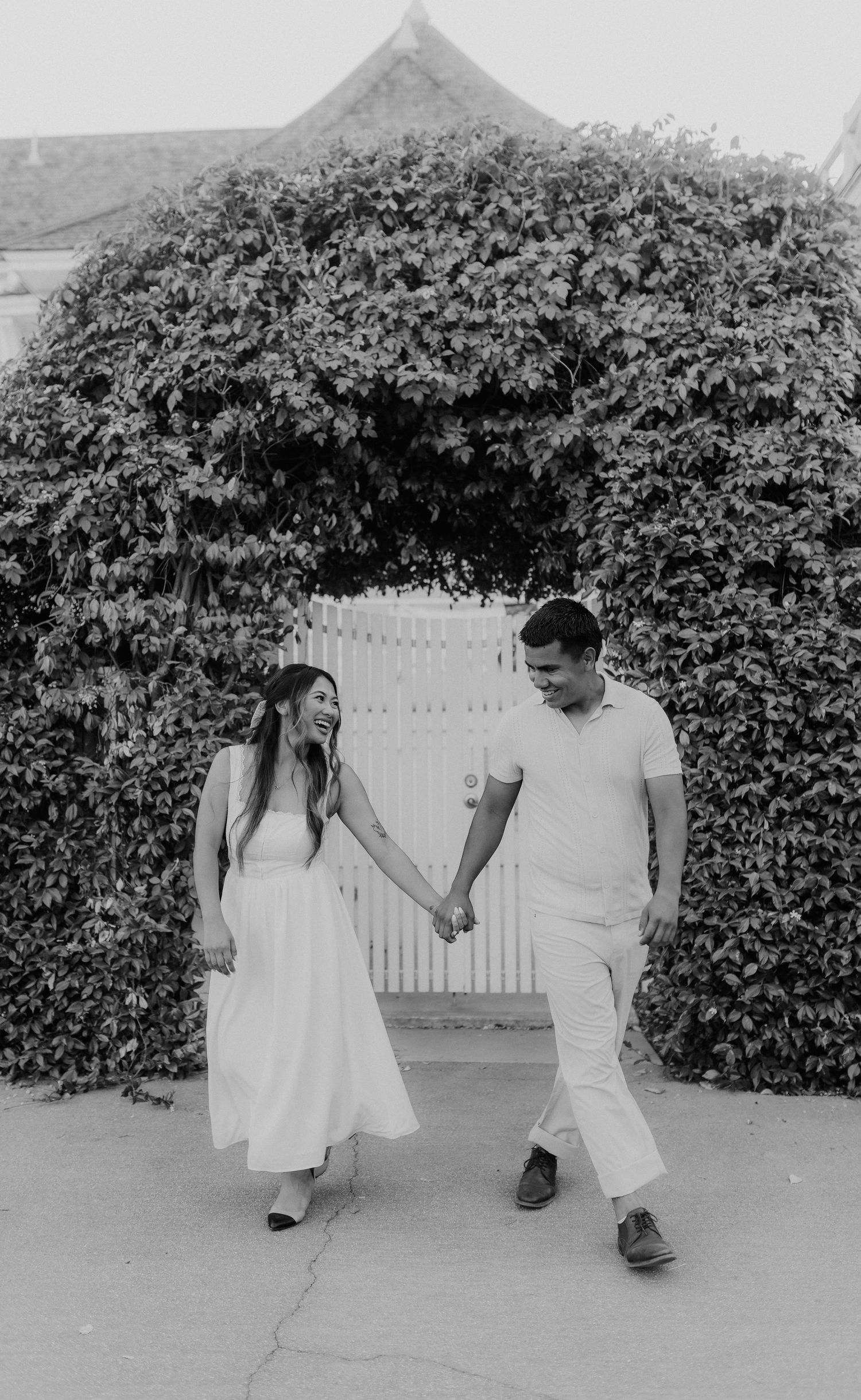 Black and white photo of couple holding hands and walking near ivy-covered gate.