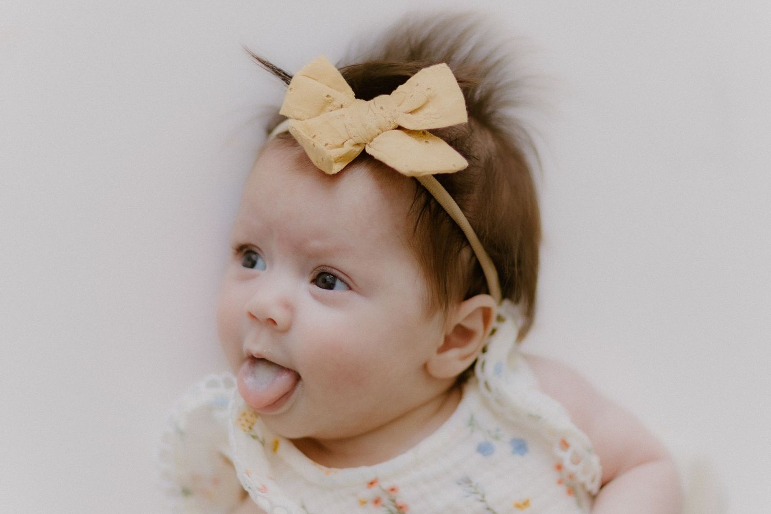Adorable baby girl with yellow bow headband sticking her tongue out playfully.