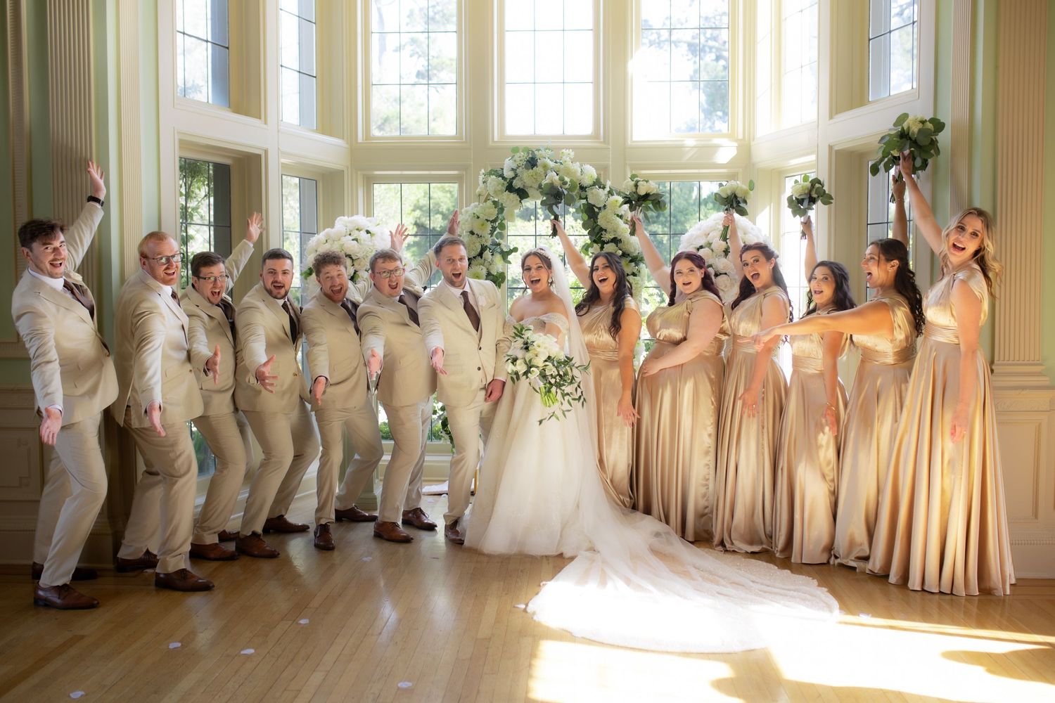 Joyful wedding party celebrates in sunlit ballroom, groomsmen in tan suits, bridesmaids in champagne gowns.