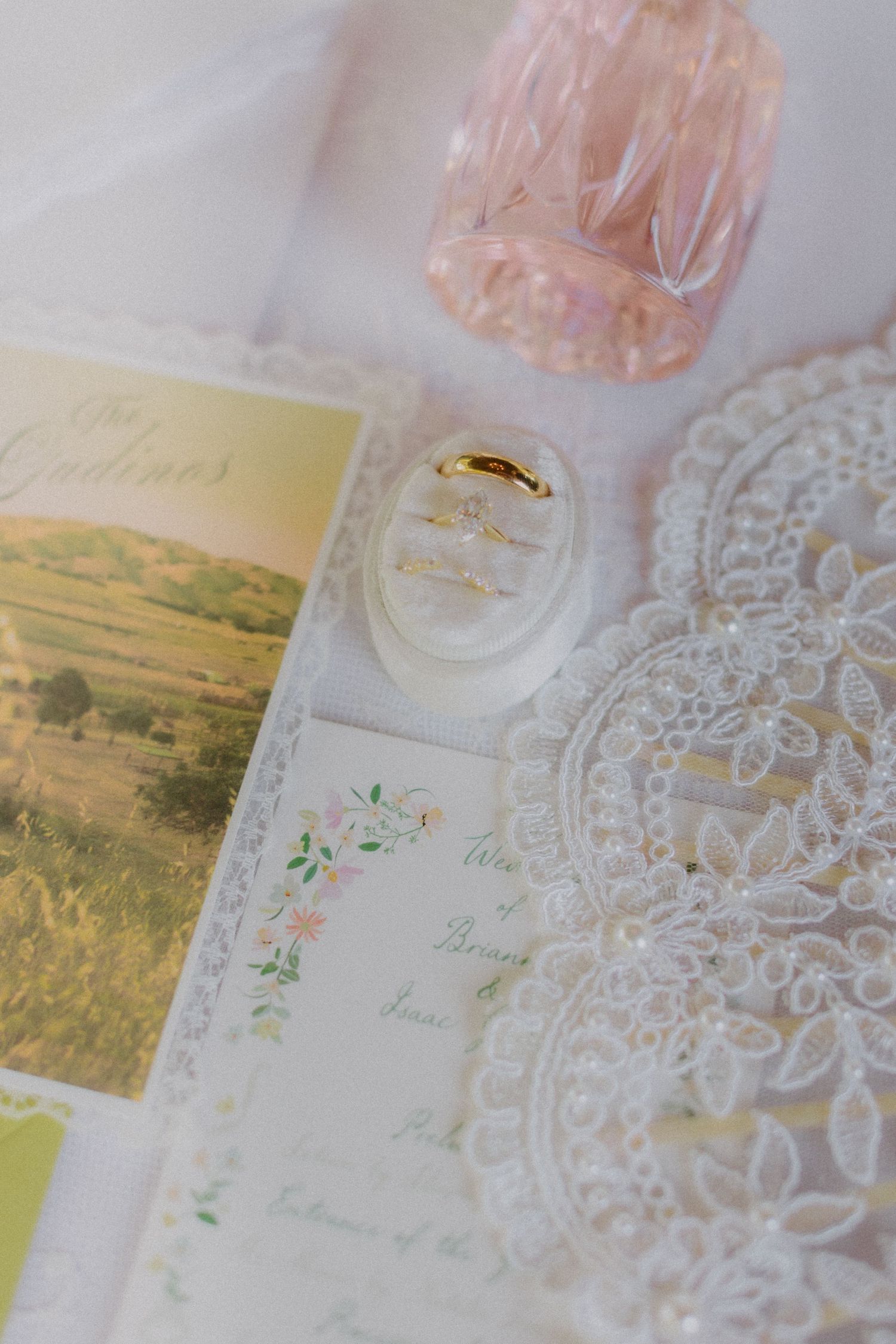 Wedding flat lay featuring gold rings, floral stationery, lace details, and a pink perfume bottle.