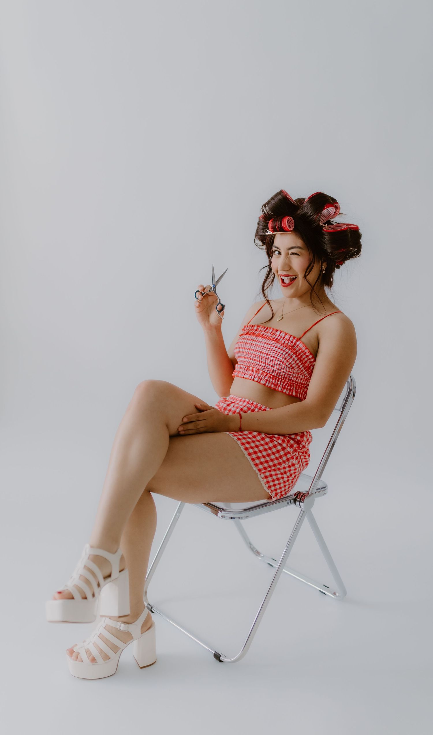 Smiling woman in red gingham outfit holding scissors sits on chrome folding chair in white studio.