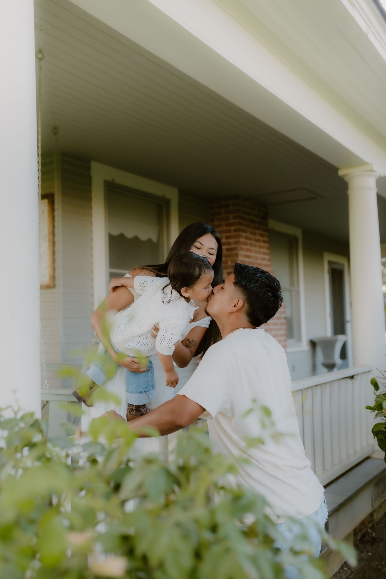 Baby girl kisses dad, mom holds her on a home porch surrounded by greenery. hanford California