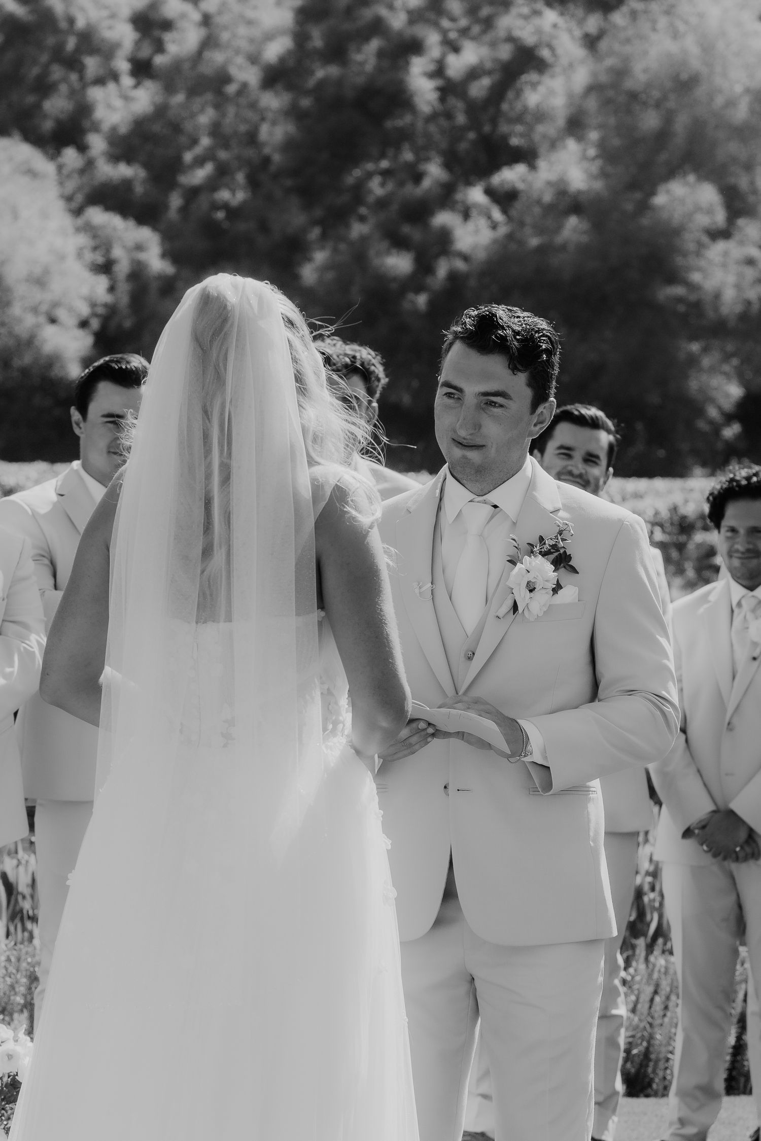Black and white photo of groom holding bride's hands during outdoor wedding ceremony with groomsmen behind.