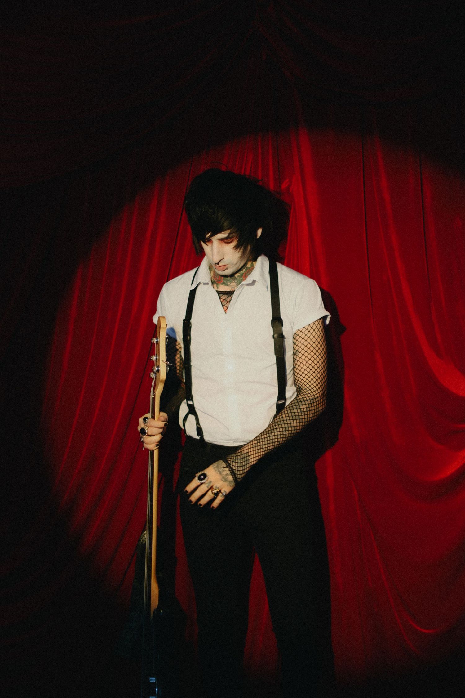 Goth musician with guitar stands in spotlight before red velvet stage curtain.