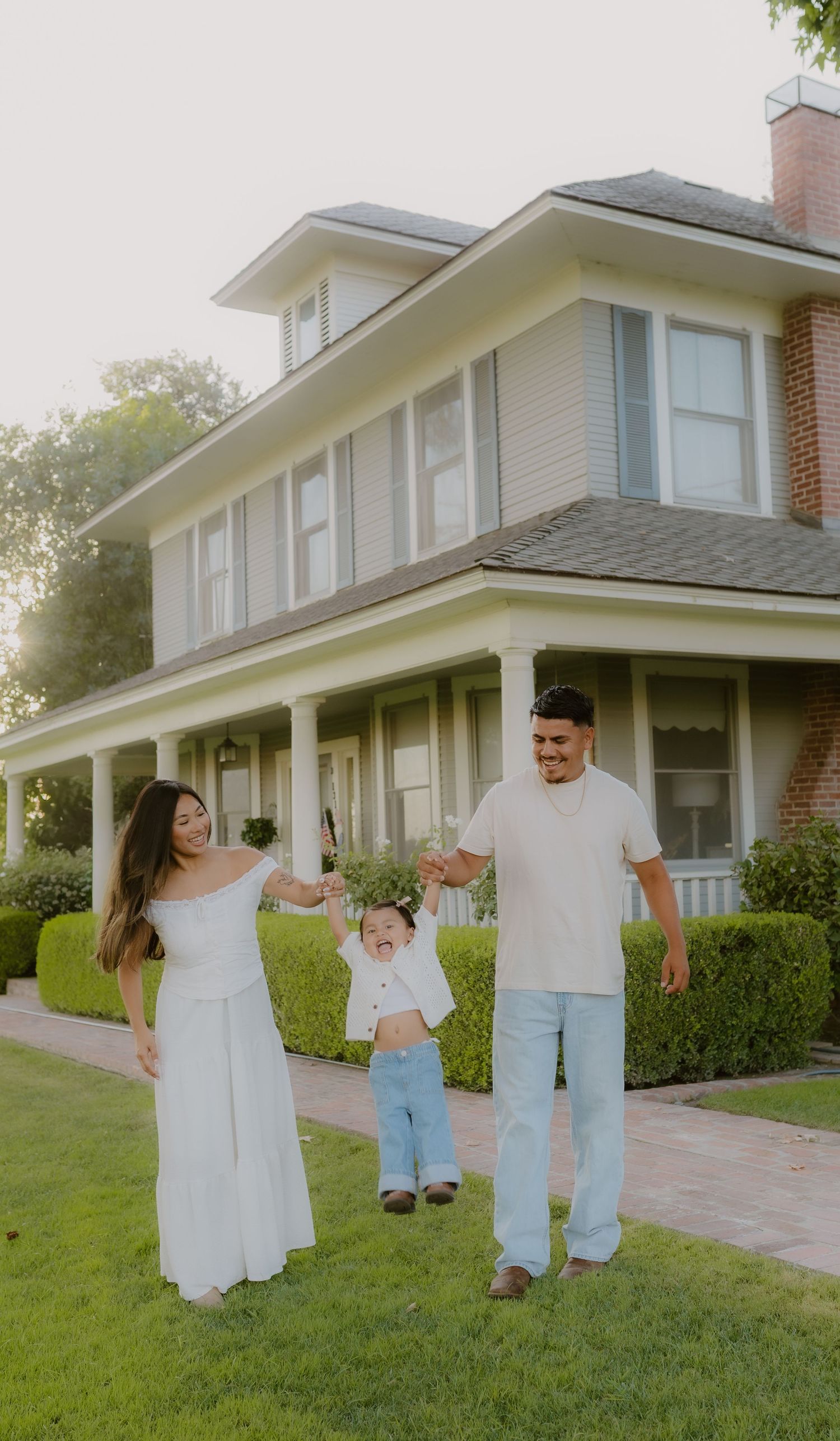 Happy family swinging toddler between parents on lawn in front of charming two-story home.