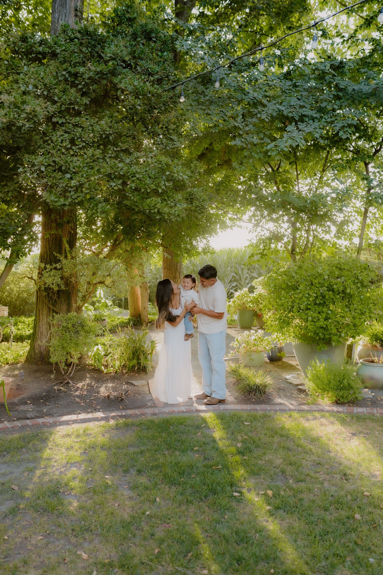 Young family with baby standing under lush green trees in sunlit garden setting.