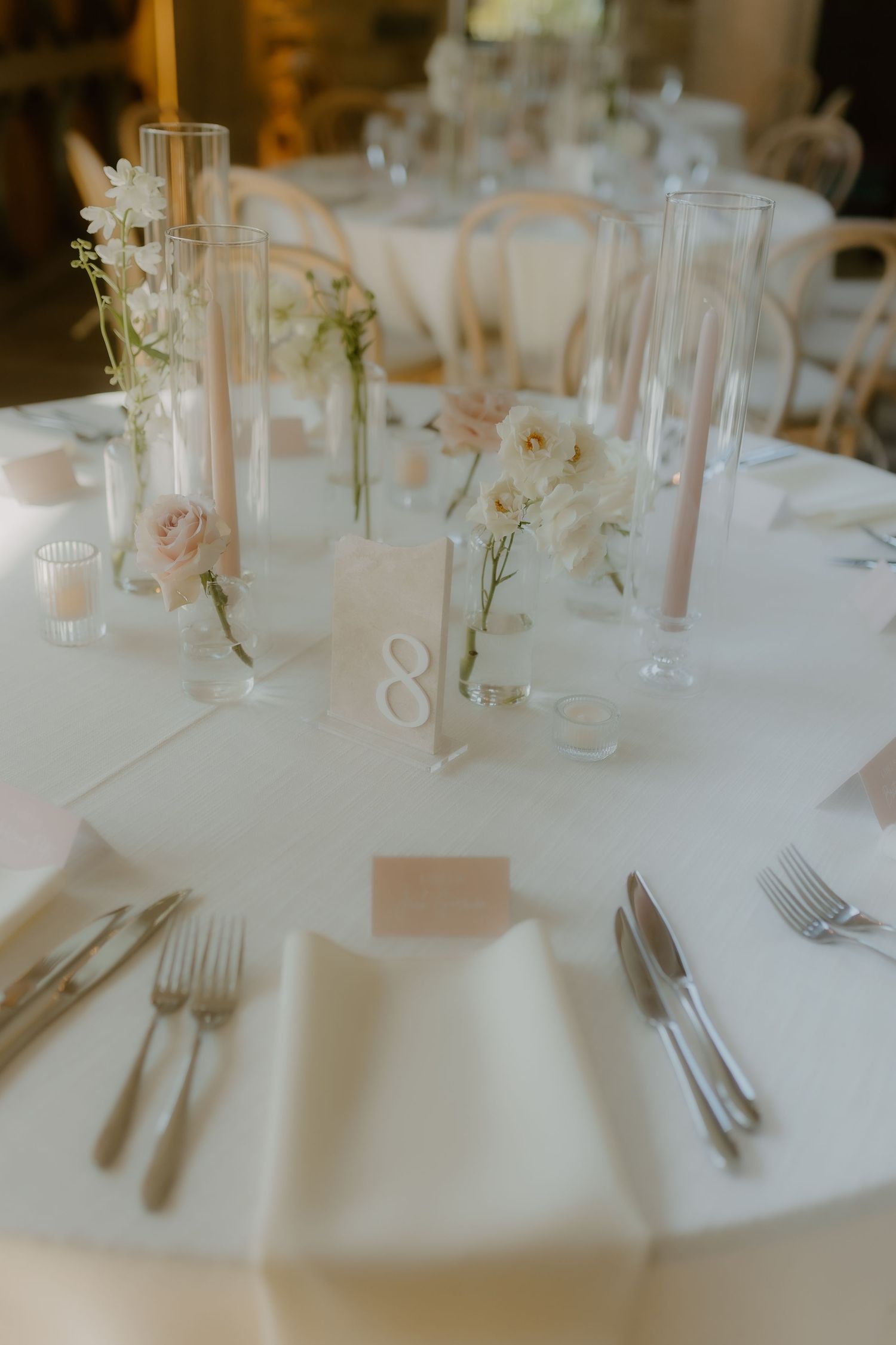 Elegant wedding reception table setting with white linens, blush roses, and tall glass vase centerpieces.