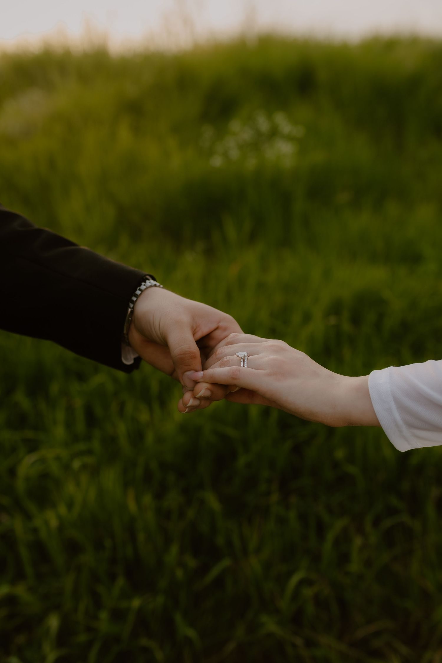 Close-up of a couple holding hands in a field, woman wearing an engagement ring, man wearing a bracelet.