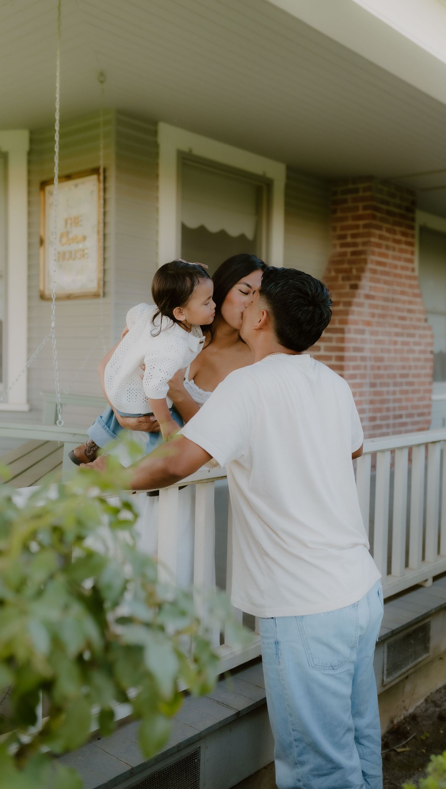 Couple sharing a kiss while holding their toddler on a white farmhouse porch at golden hour.
