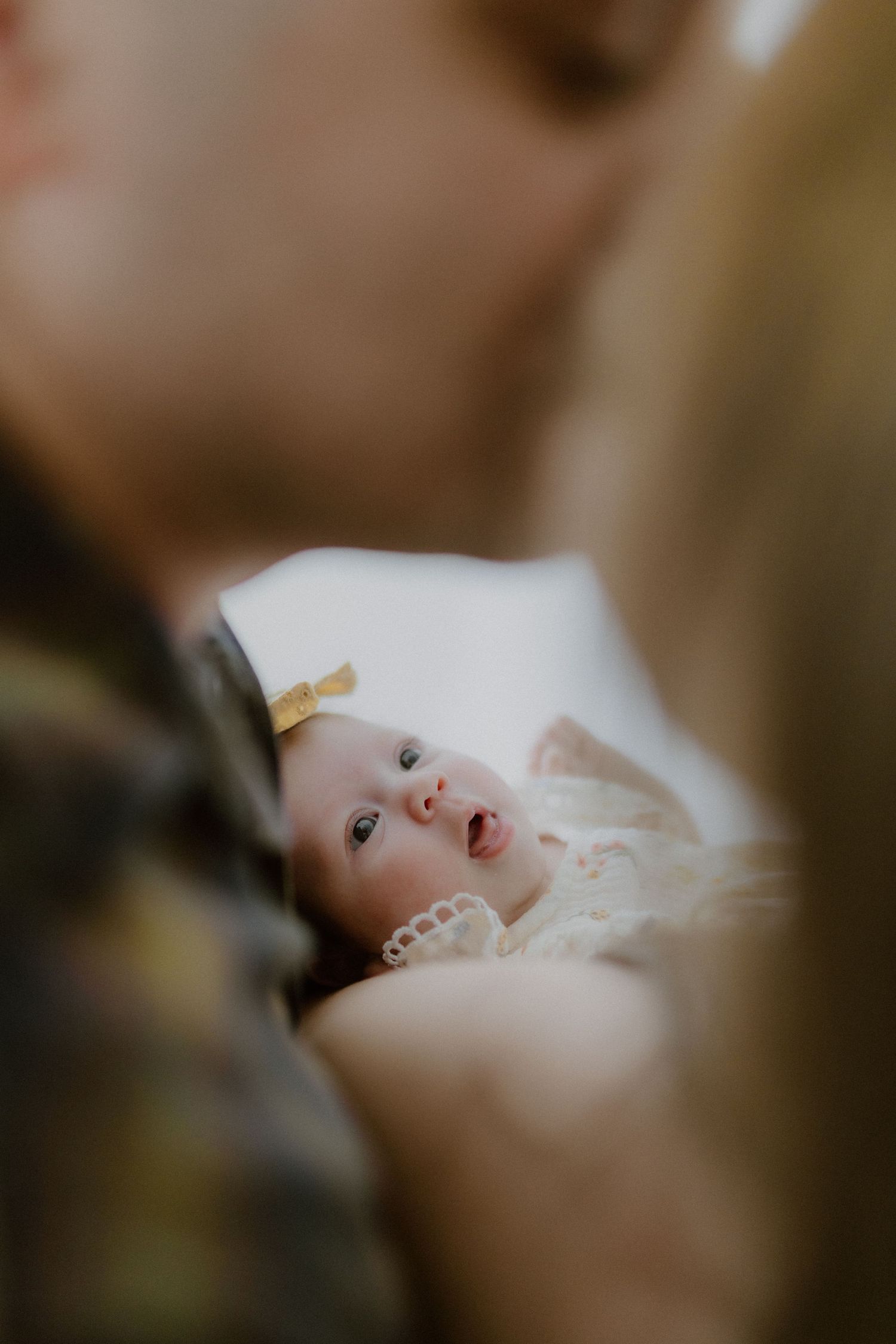 A newborn baby girl with yellow bow looks up while cradled between her parents in warm tones.
