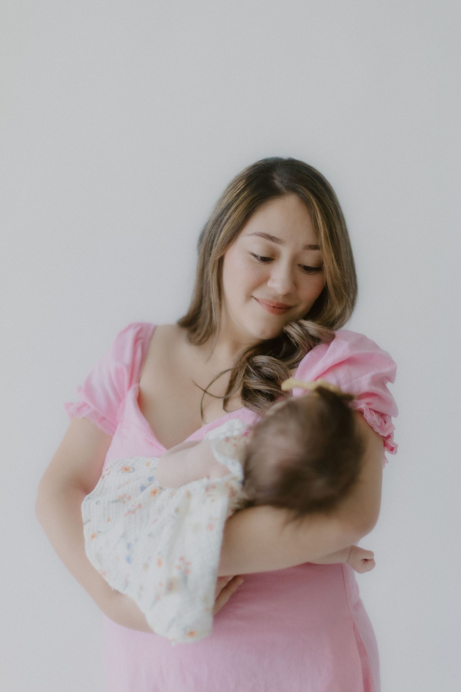 A mother in a pink dress smiles lovingly while holding her swaddled newborn baby.