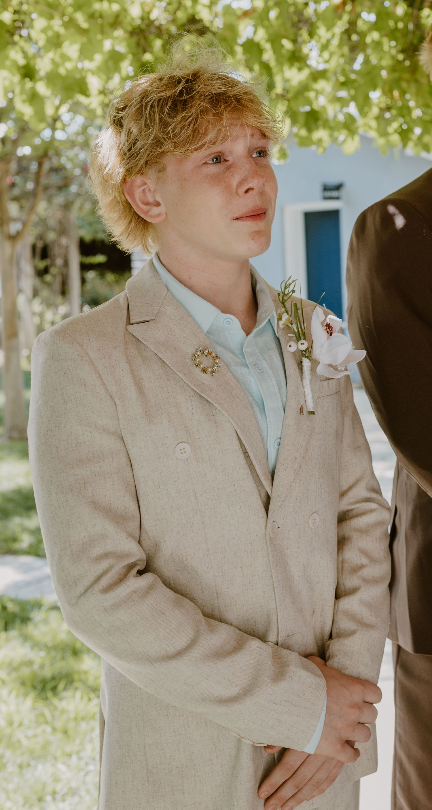 Emotional young boy in beige suit with boutonniere crying at outdoor wedding ceremony.