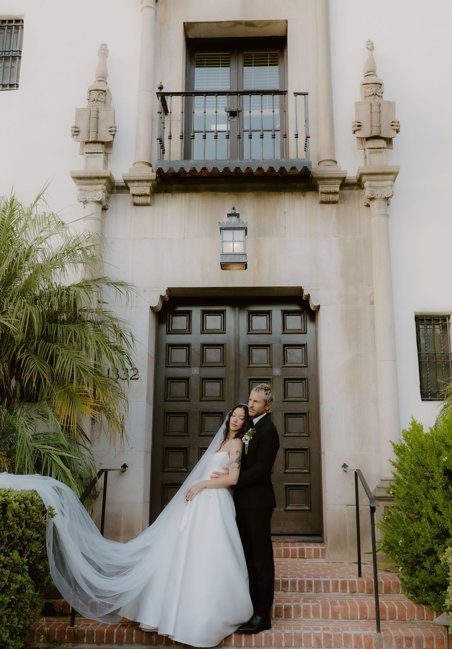 Bride's veil flows dramatically as couple embraces before grand dark doors of a Spanish Colonial building.