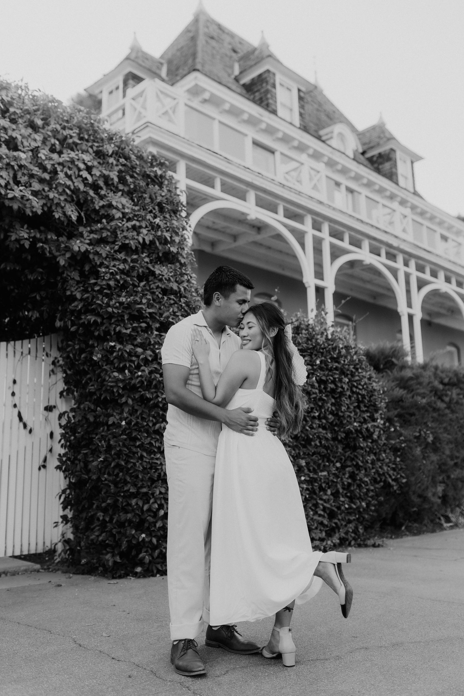 Black and white photo of couple dancing joyfully outside an ivy-covered Victorian estate.