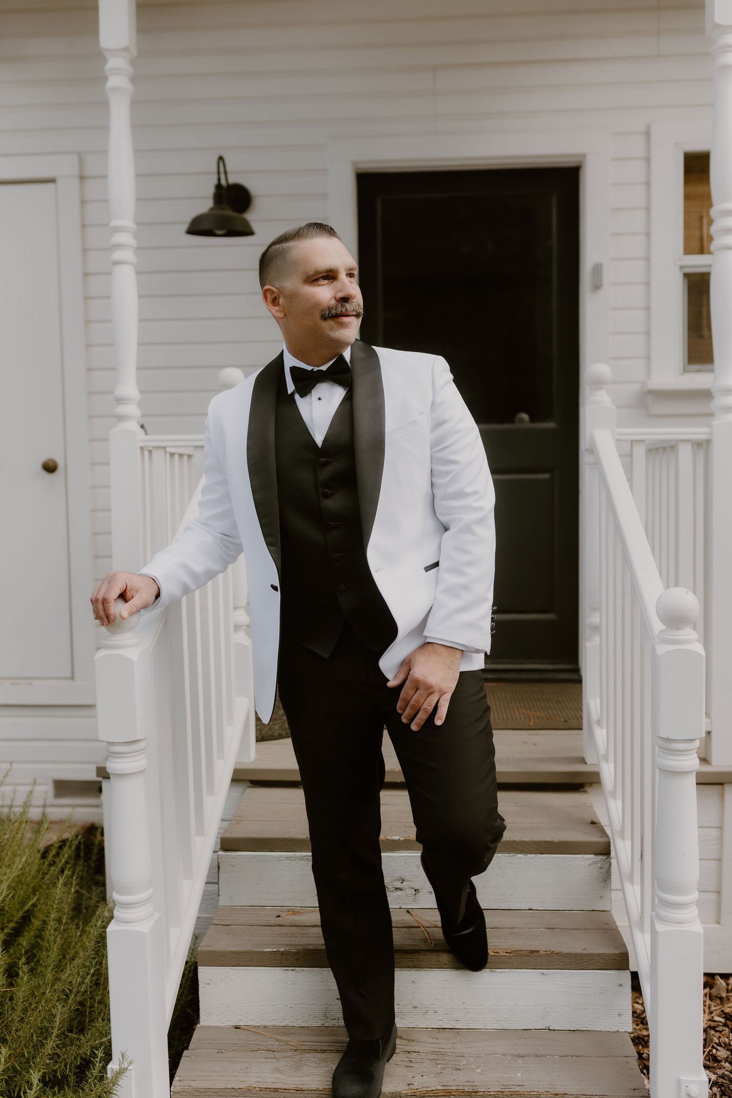 Dapper groom in white tuxedo jacket and black bow tie posing on steps of white wooden building.