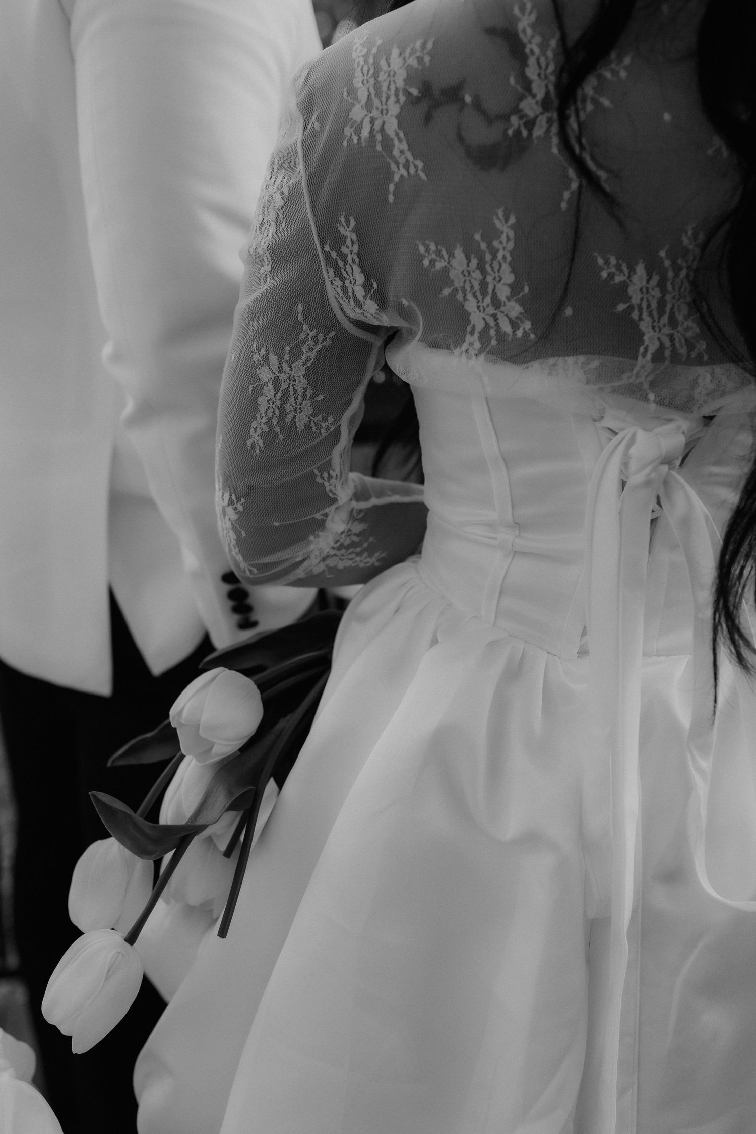 Black and white close-up of bride in lace corset gown holding tulips beside groom in white jacket.