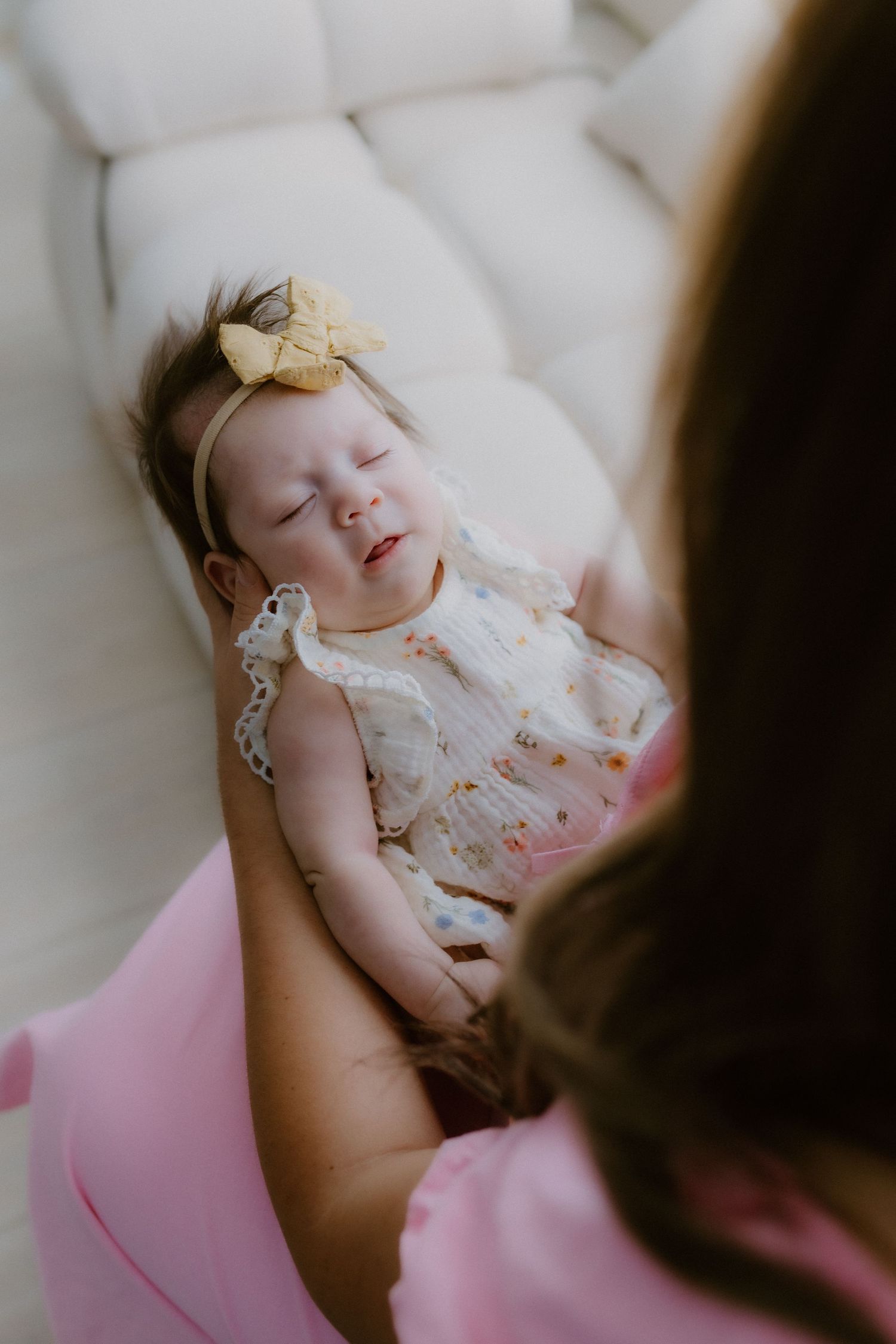 Overhead view of a baby girl in a floral outfit and yellow bow headband smiling while held by her mother.