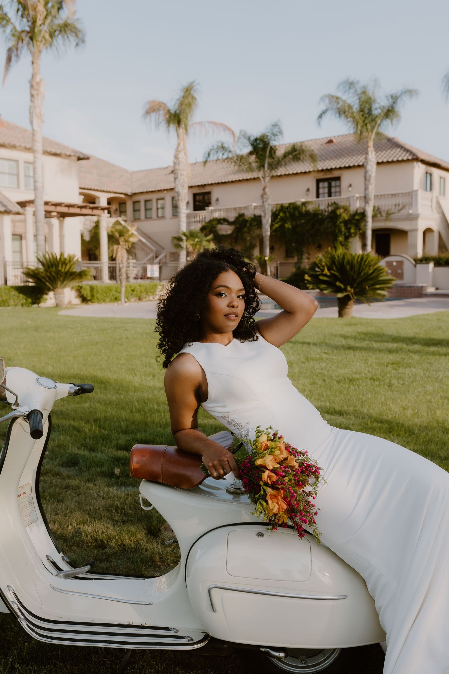 Bride in sleek white gown poses on white Vespa scooter holding colorful bouquet at sunset. Vista Del Lago Estate, Bakersfield, cinematic