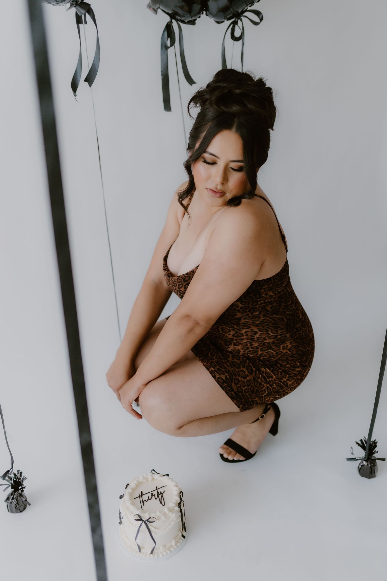 Woman in leopard print dress crouching beside small cake with black balloons in white studio. birthday, Tulare California