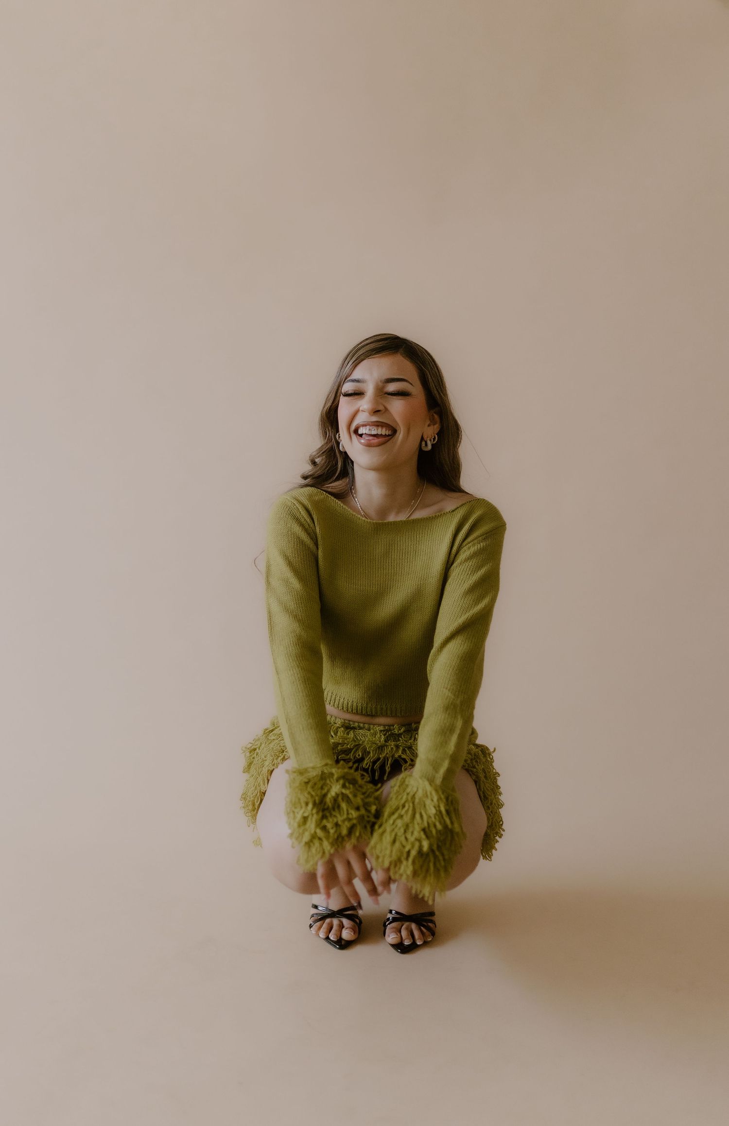 Laughing woman in olive green long-sleeve crop top and feathered mini skirt crouches on beige backdrop.