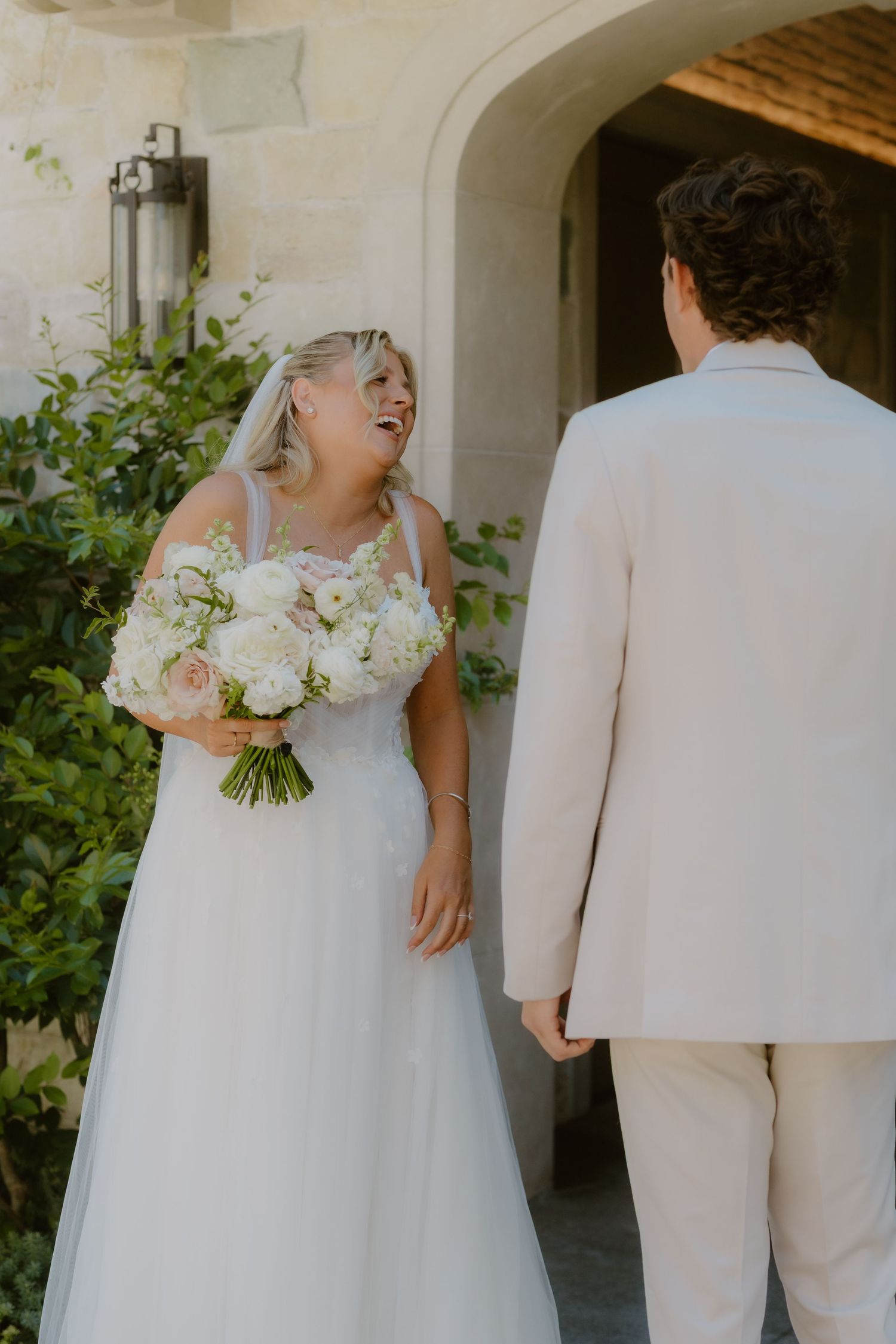 Smiling bride in a tulle gown holding white bouquet during first look with groom outside stone venue.