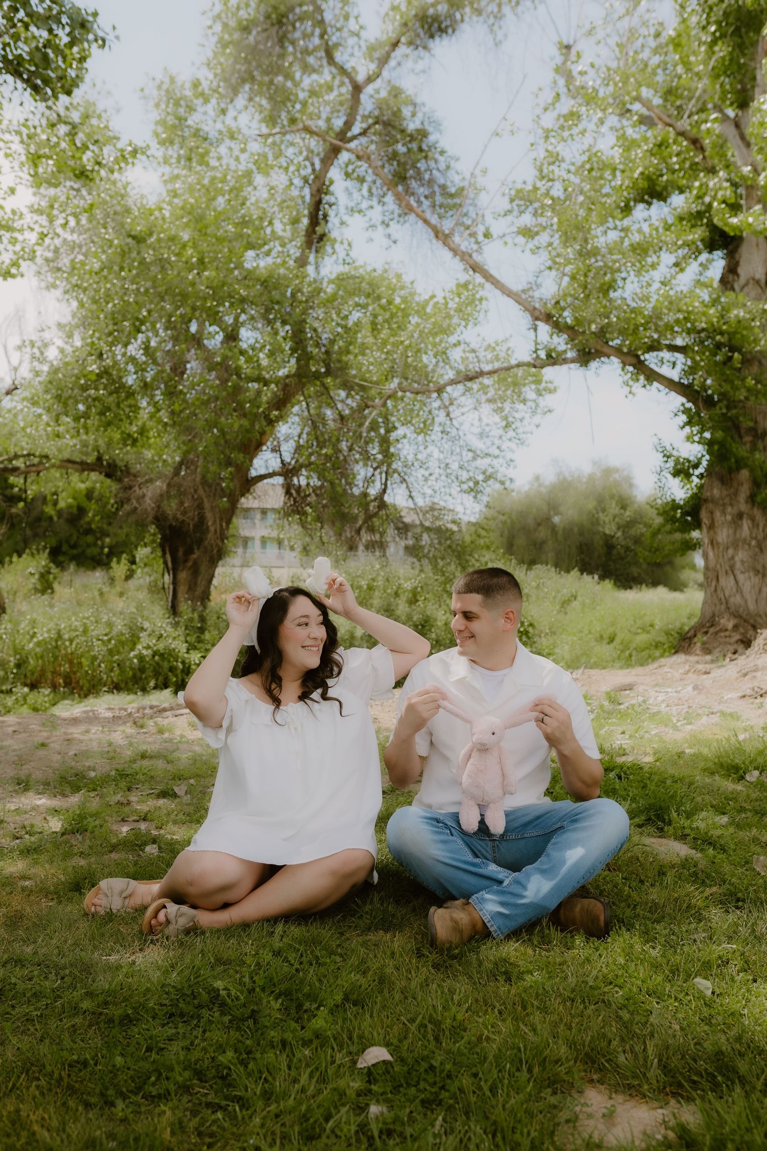 Couple sitting on grass under trees, man holding baby shoes while woman raises hands joyfully during maternity photo session.