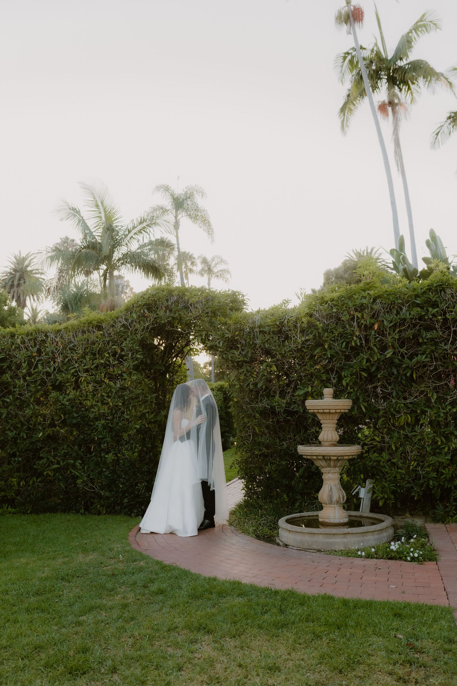 Bride and groom share an intimate veiled moment near a stone fountain surrounded by tall green hedges and palms.