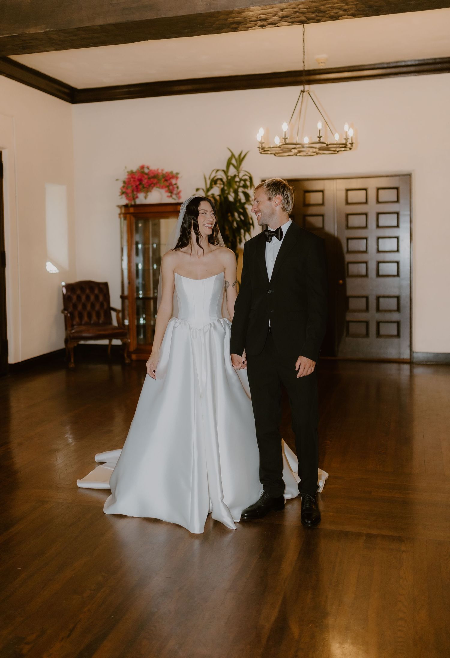 Bride and groom in elegant wedding attire hold hands in a classic wood-paneled reception hall.