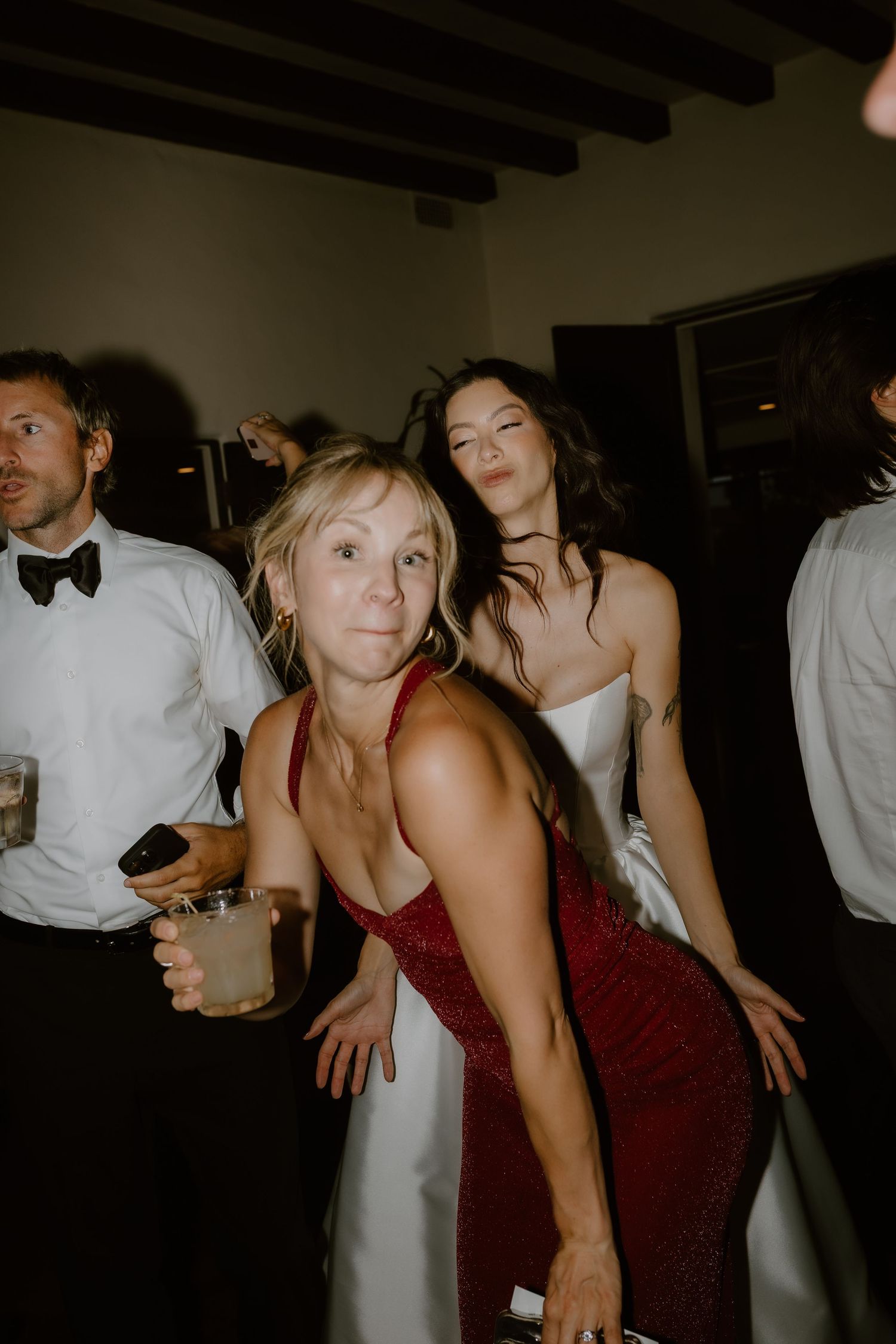 Bride in white gown dancing with guest in red dress at lively wedding reception party.