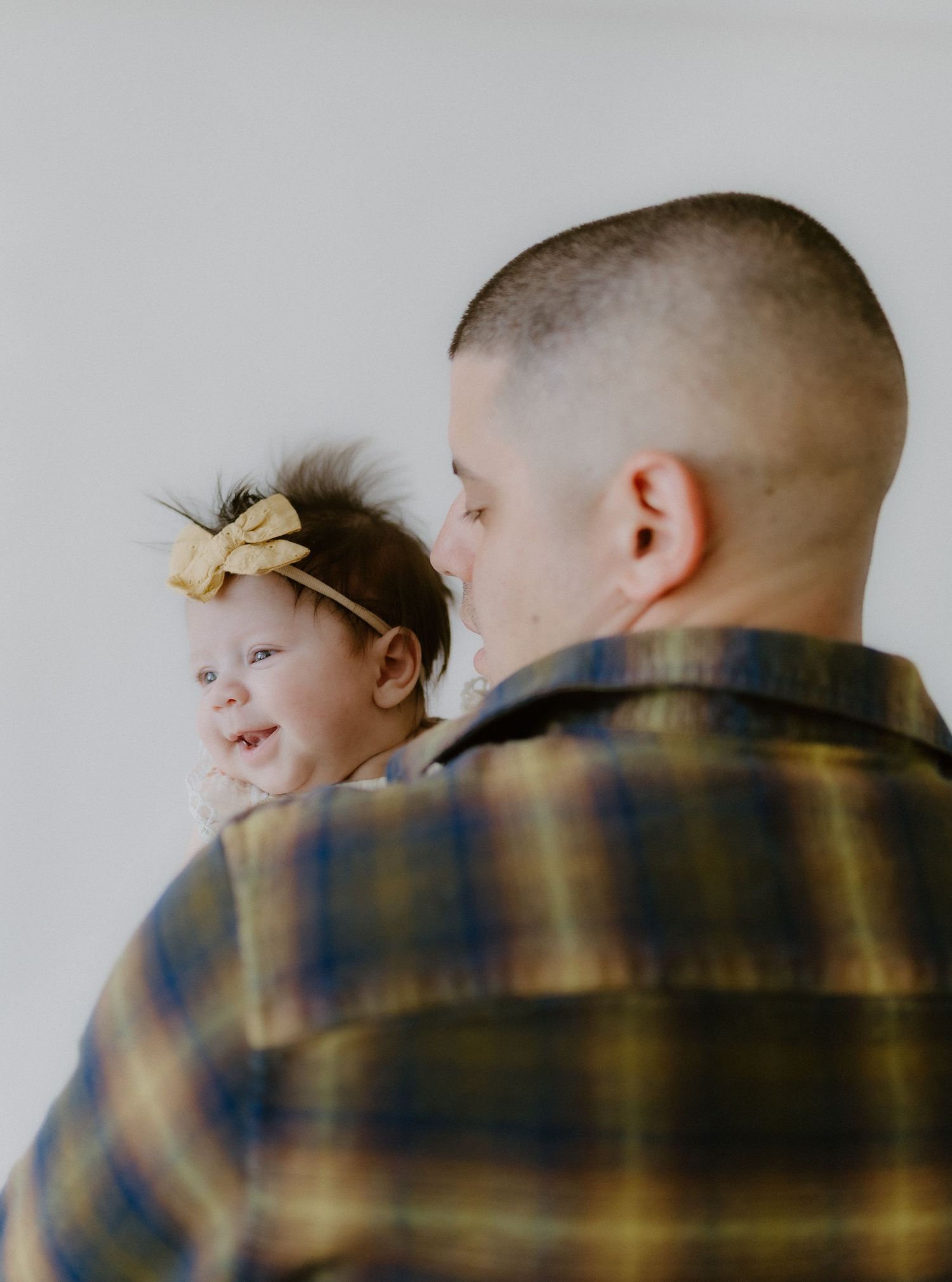A father in yellow plaid shirt holds his smiling newborn baby girl with yellow bow headband.