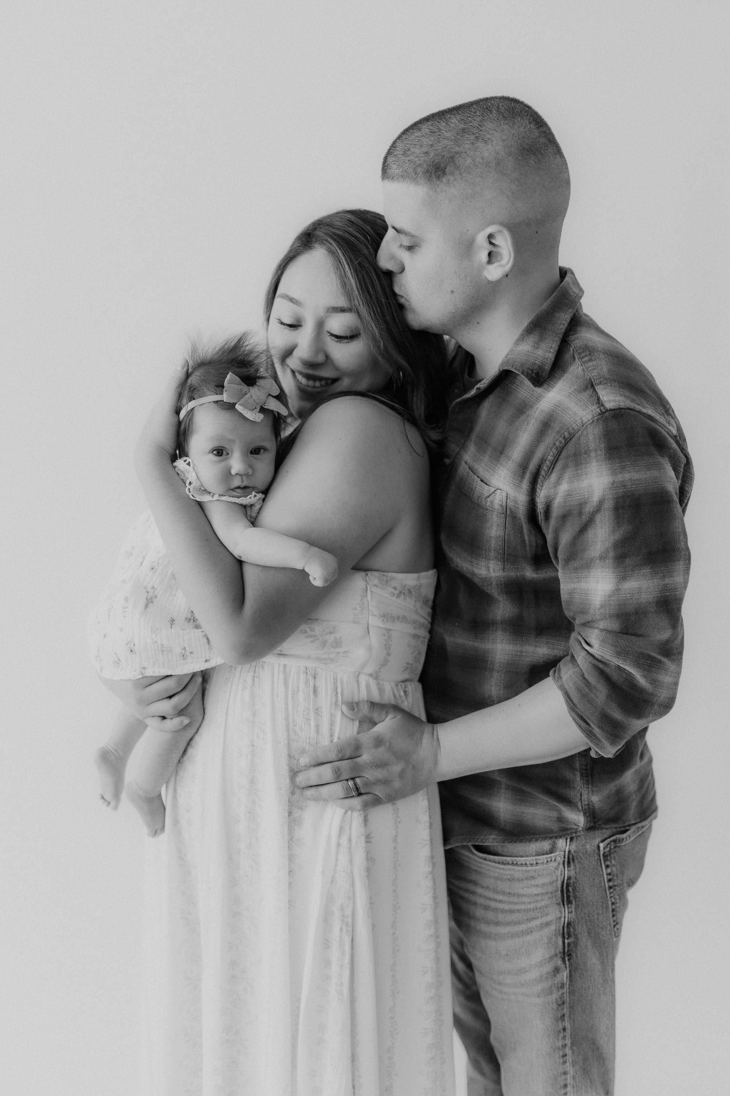 Black and white family portrait of parents with newborn, father kissing mothers temple as she holds baby.