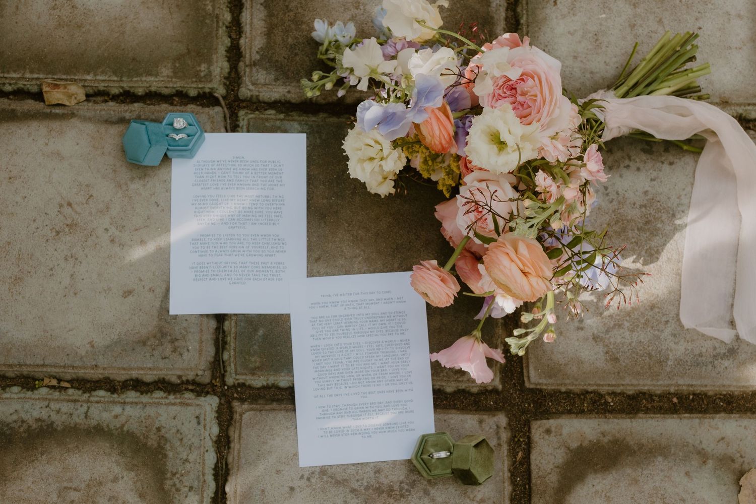 Wedding flat lay featuring pastel bridal bouquet, vow letters, and turquoise ring box on stone.
