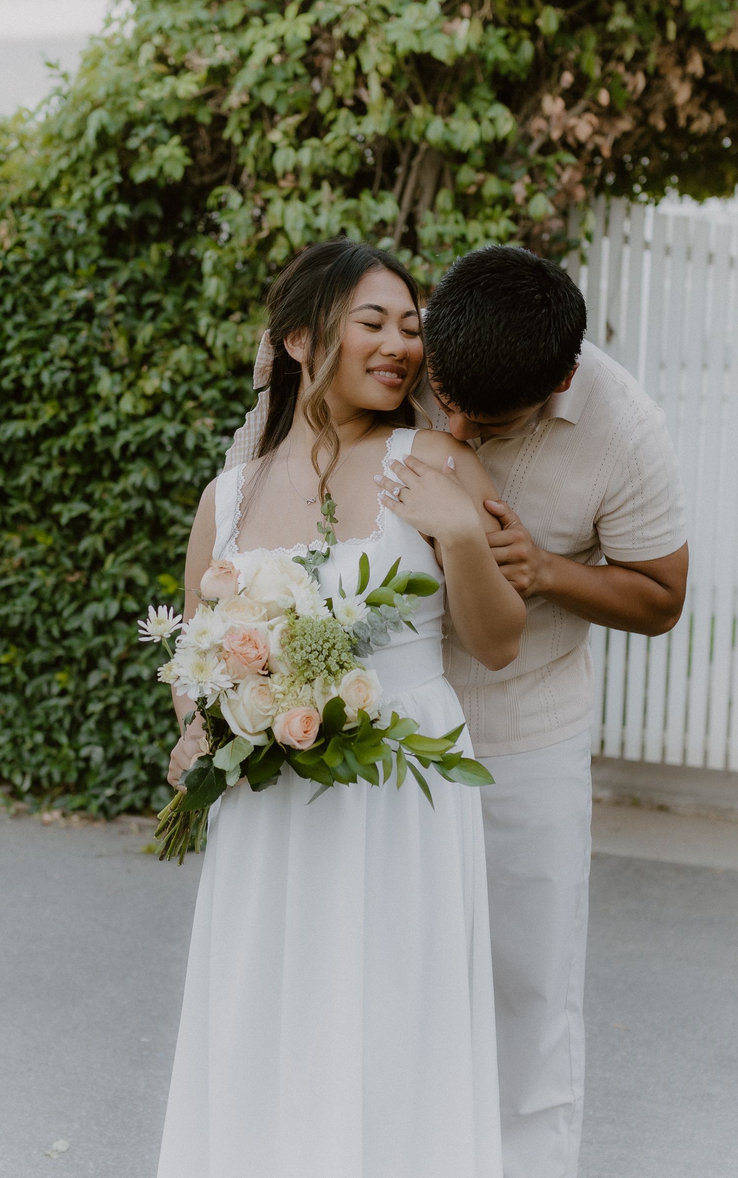 Smiling bride holding a peach and white bouquet while groom embraces her from behind outdoors.