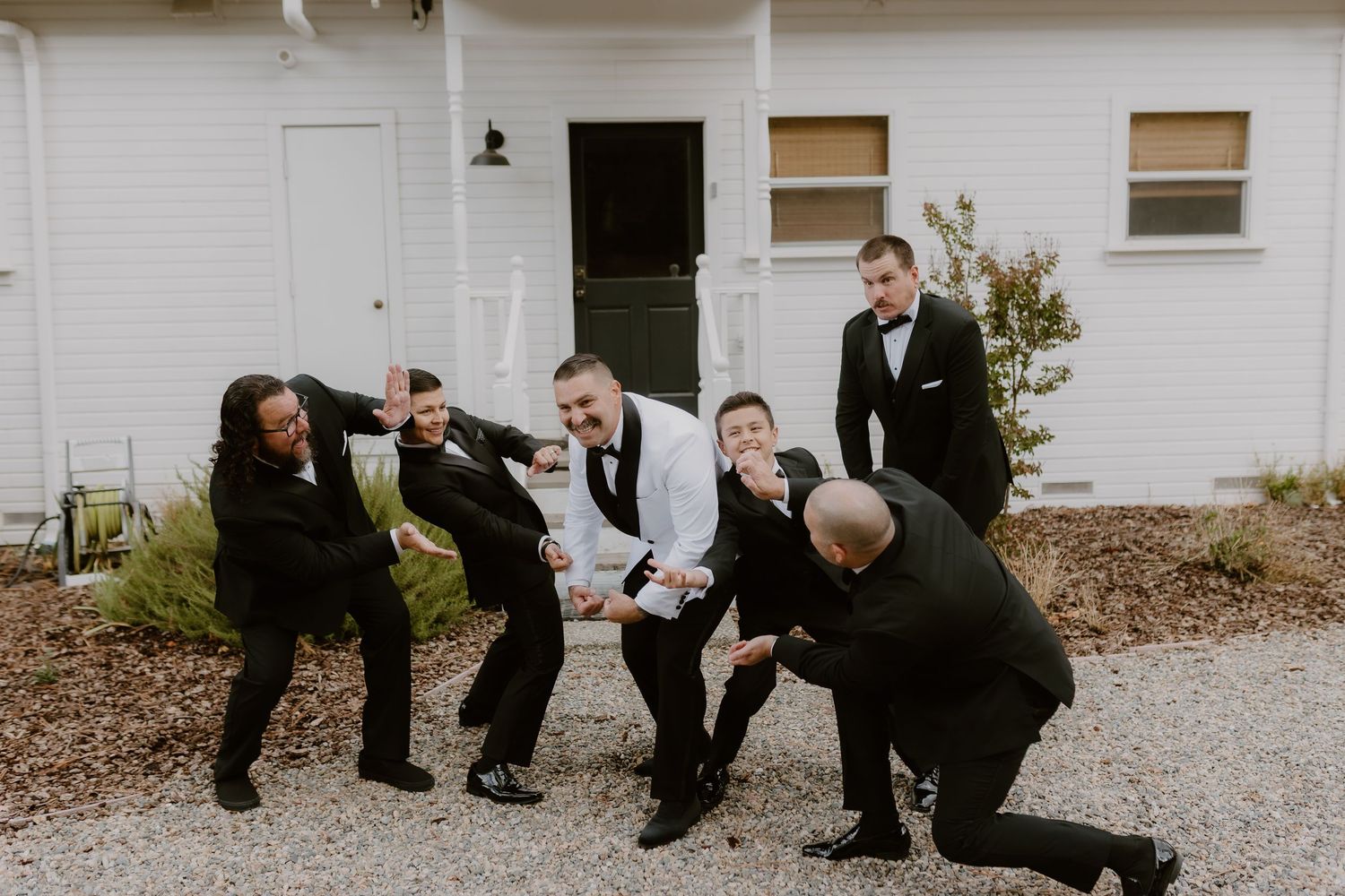 Groom and groomsmen in black tuxedos celebrating with champagne outside white building in fun candid photo.