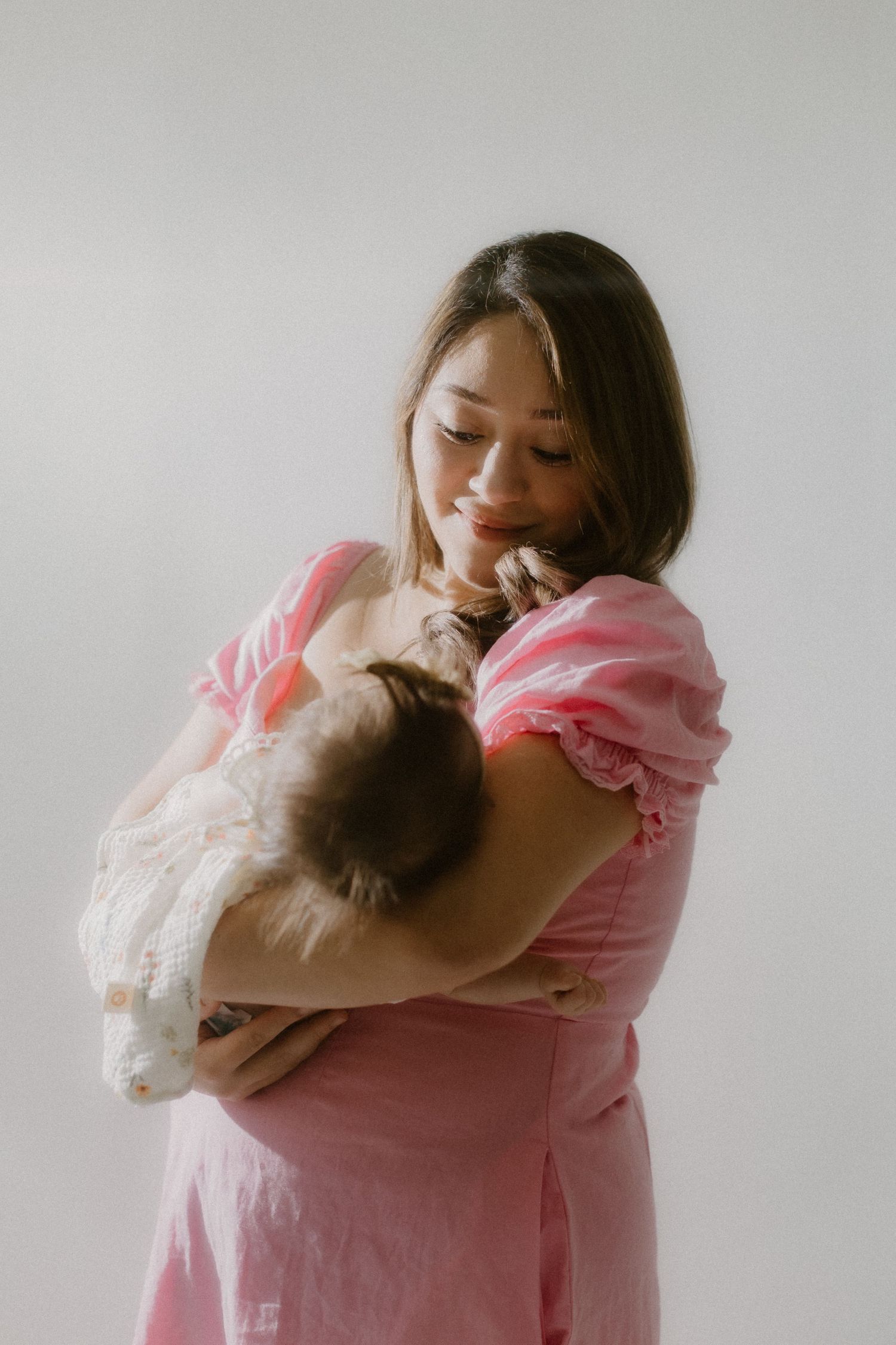 Young mother in pink dress standing against white wall gently holding and gazing at newborn baby.