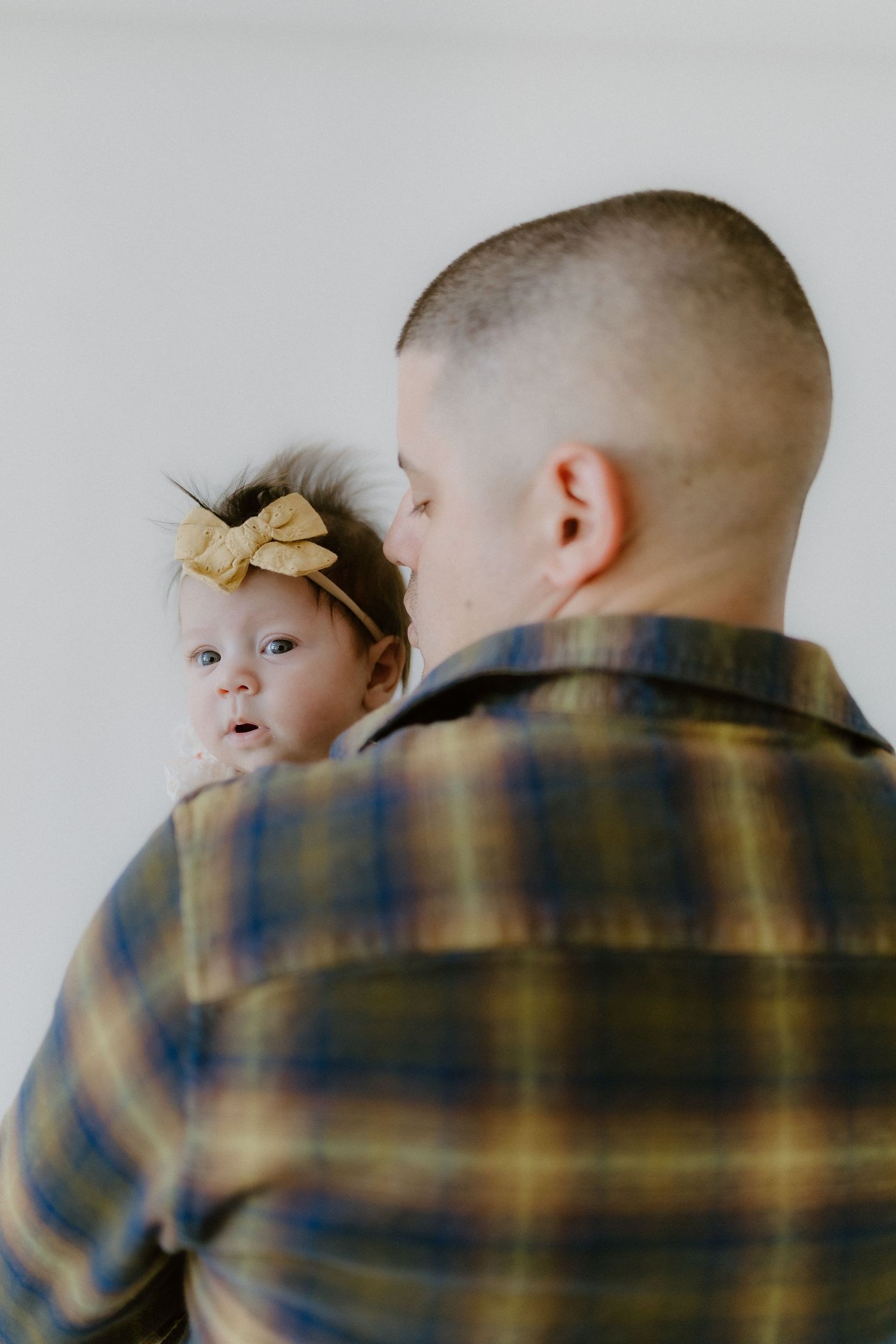 A father in plaid shirt holds his wide-eyed newborn baby girl wearing a yellow bow headband.