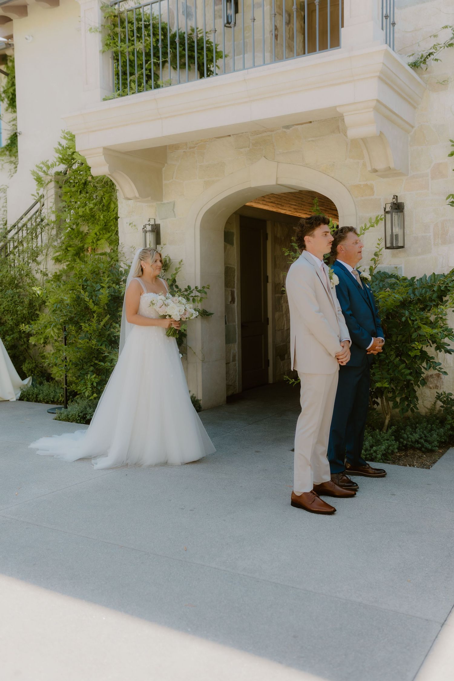 Bride in white gown holds bouquet during first look moment with groom in tan suit outdoors.