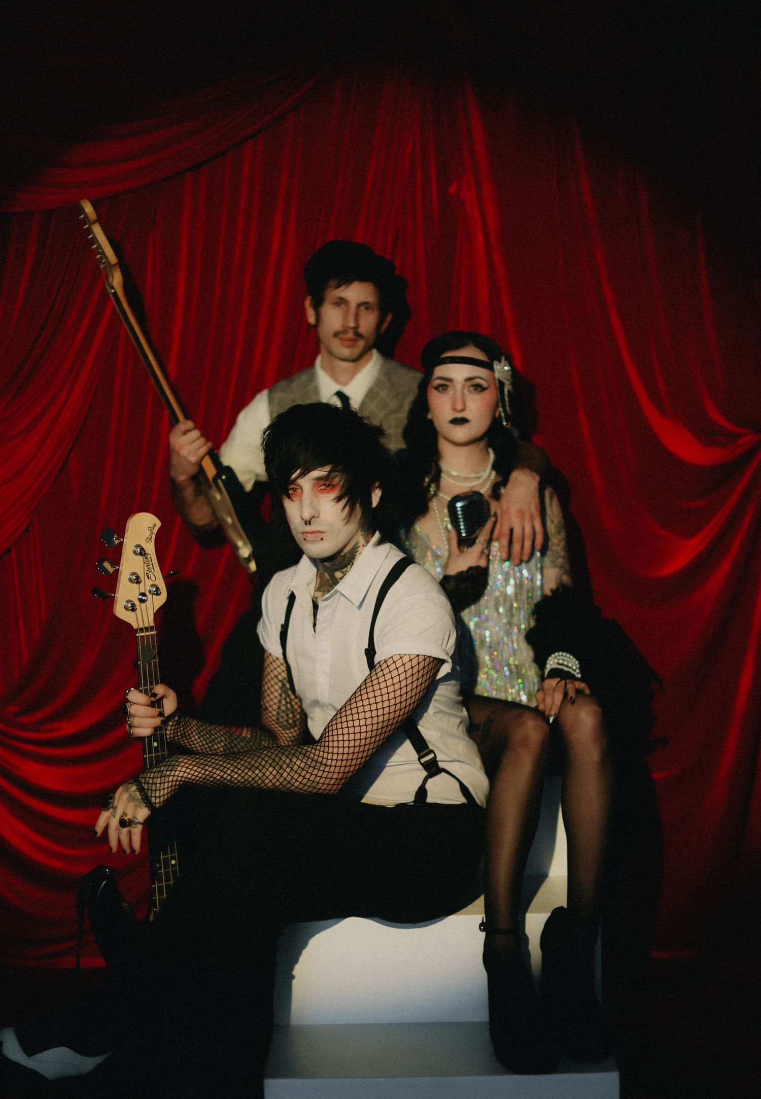 Three performers in vintage cabaret costumes pose dramatically before red velvet curtains.
