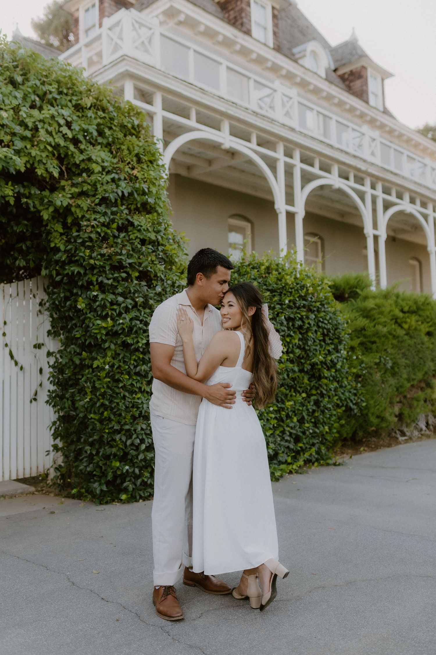 Romantic couple in white outfits embracing in front of ivy-covered Victorian building.