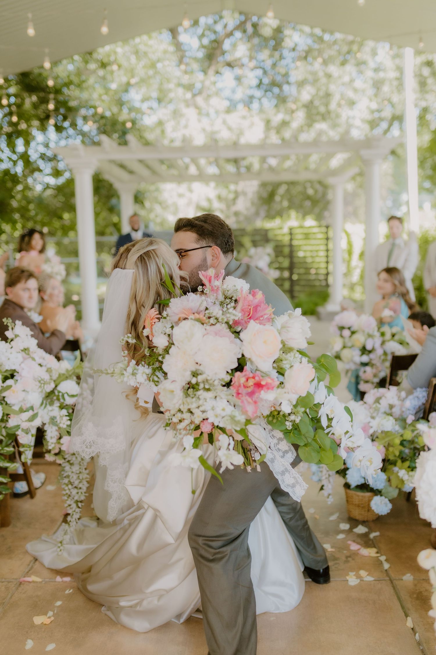 Newlyweds sharing romantic kiss with large pink and white floral bouquet at wedding ceremony.